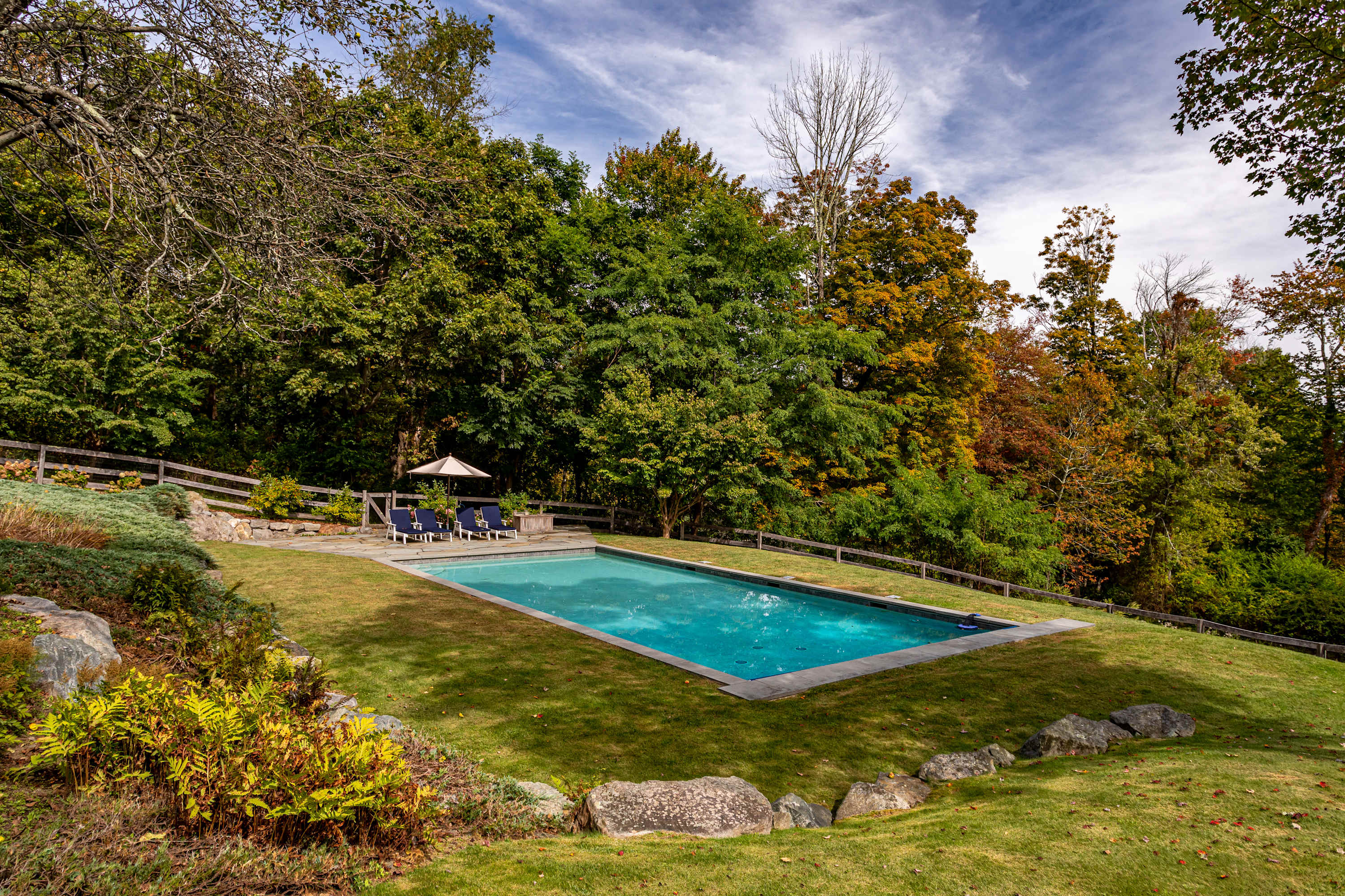 An inviting in-ground rectangular swimming pool with bright blue water is nestled into a lush grassy hillside, surrounded by a vibrant mix of green and autumn-colored trees under a partly cloudy sky. A stone patio with lounge chairs and an umbrella offers a relaxing spot beside the pool.