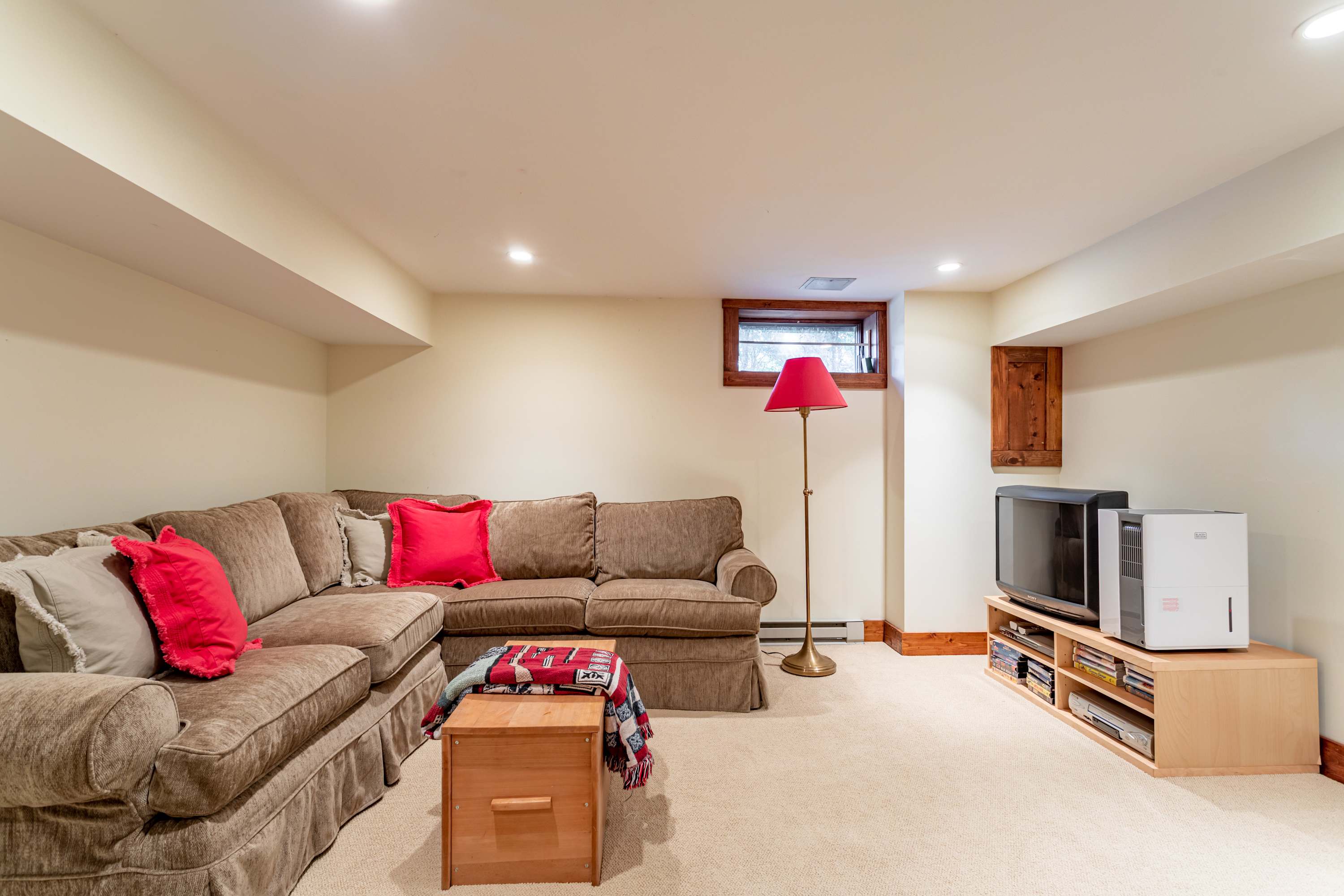 Cozy corner of a recreation room featuring a large sectional sofa with red accent pillows, facing a small TV setup. A floor lamp provides ambient lighting, and a small window is visible above.