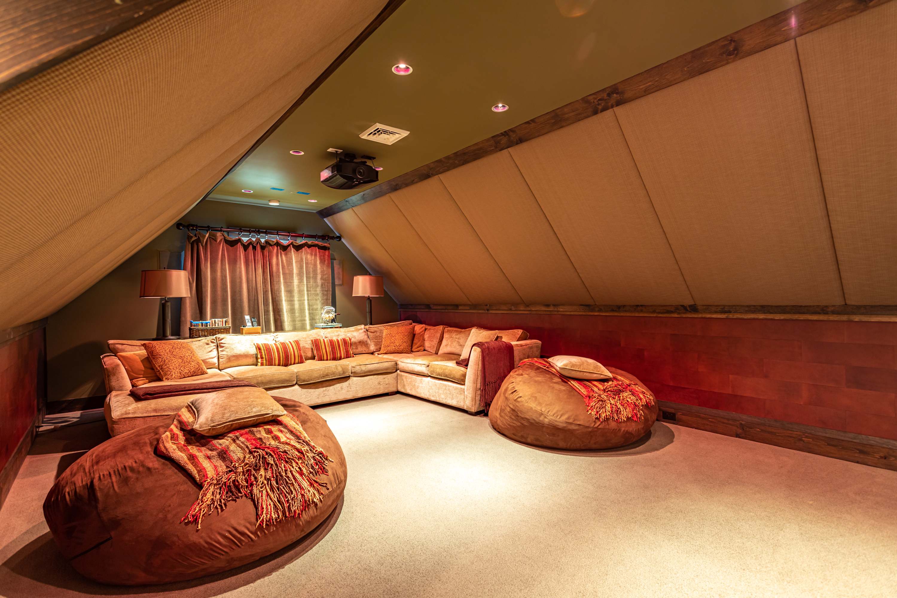 Cozy media room with a large L-shaped sectional sofa and two bean bag chairs, set under a sloped ceiling with sound-absorbing panels. A projector is mounted on the ceiling, creating an intimate viewing experience.