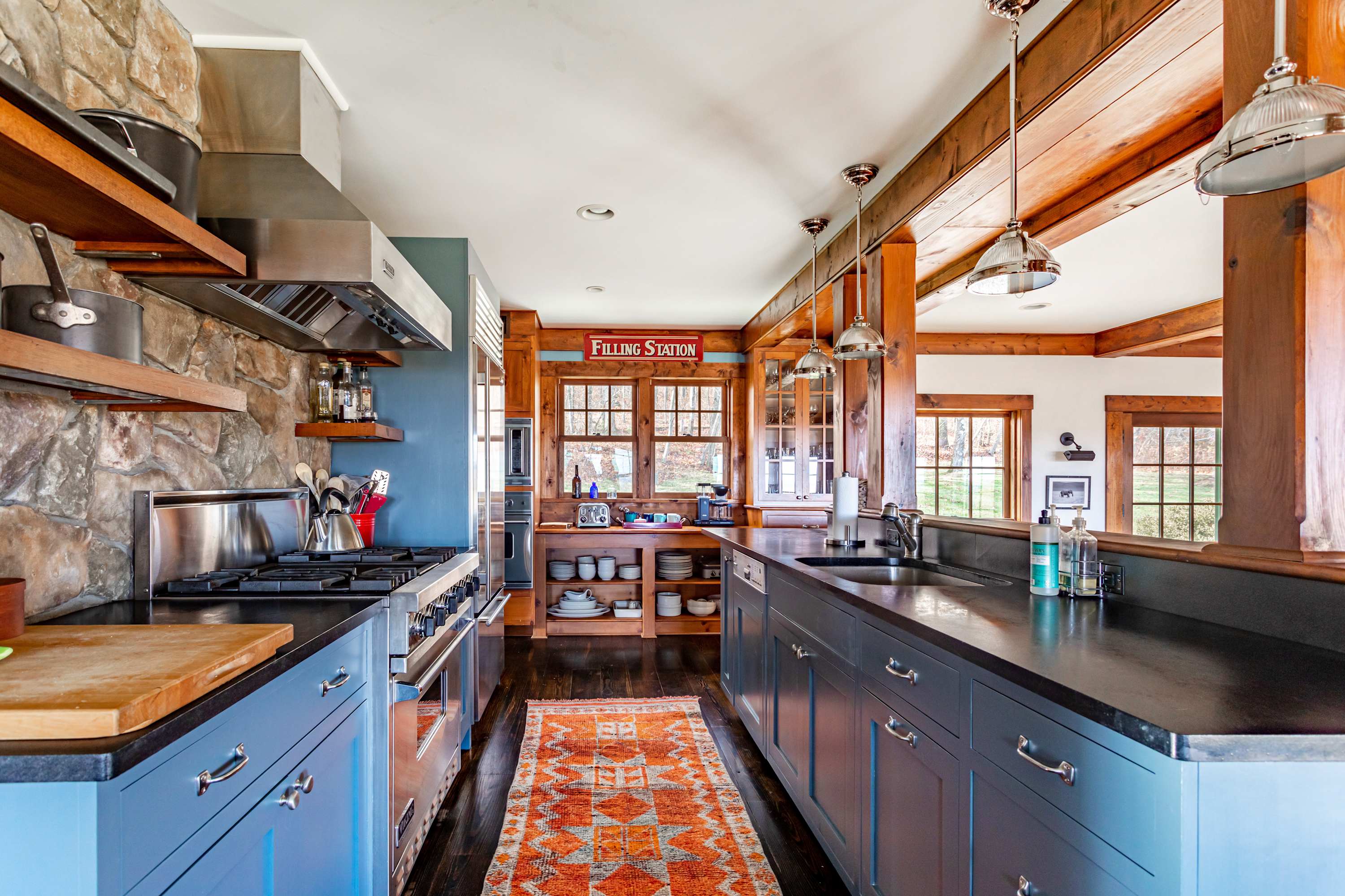A rustic yet modern kitchen features blue cabinetry, dark countertops, a stainless steel range with a stone backsplash, and exposed wooden beams, with an orange patterned runner rug.