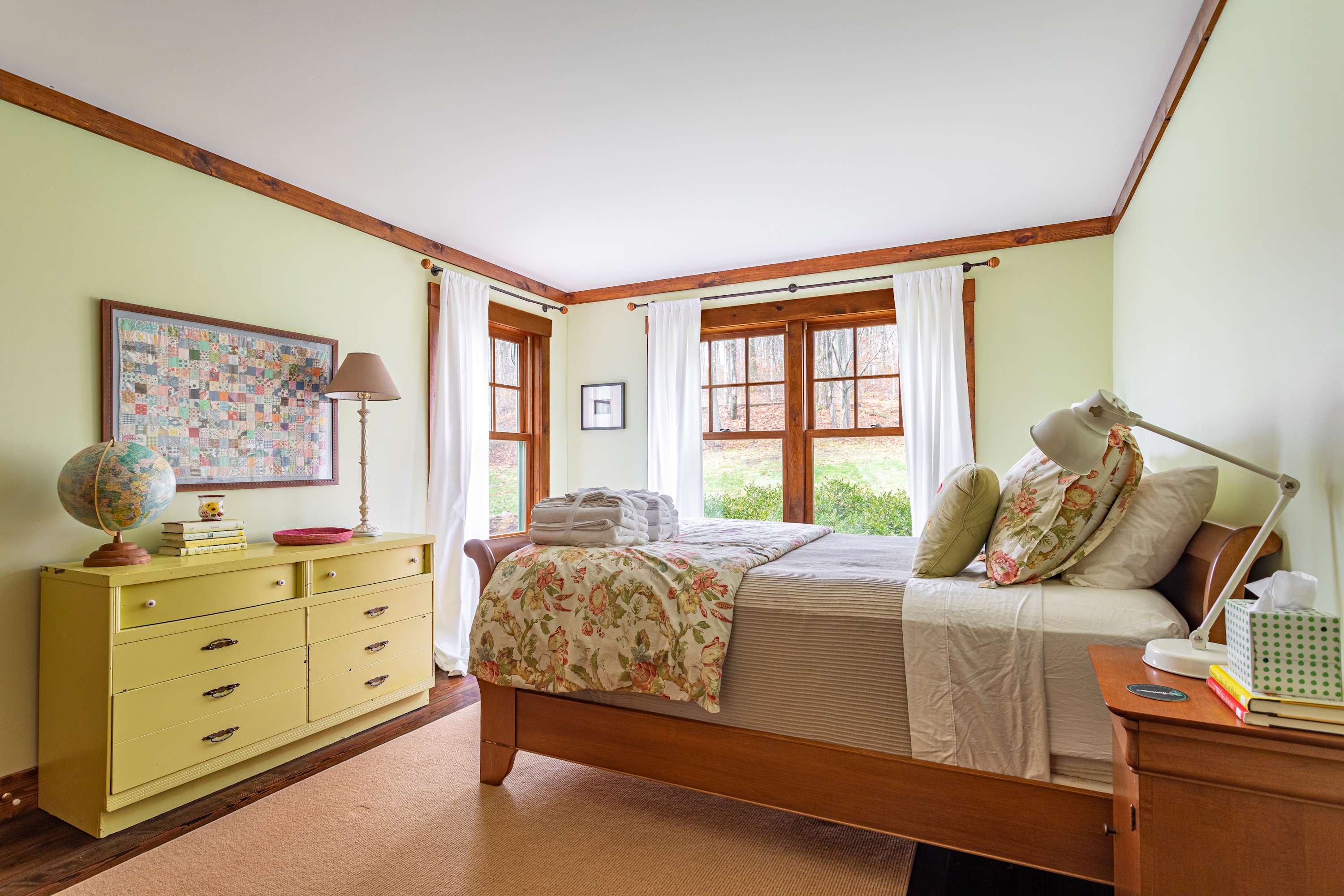 A cozy bedroom with light green walls, a wooden sleigh bed dressed in floral and striped bedding, a yellow dresser with a globe and lamp, and two large windows overlooking a wooded area.