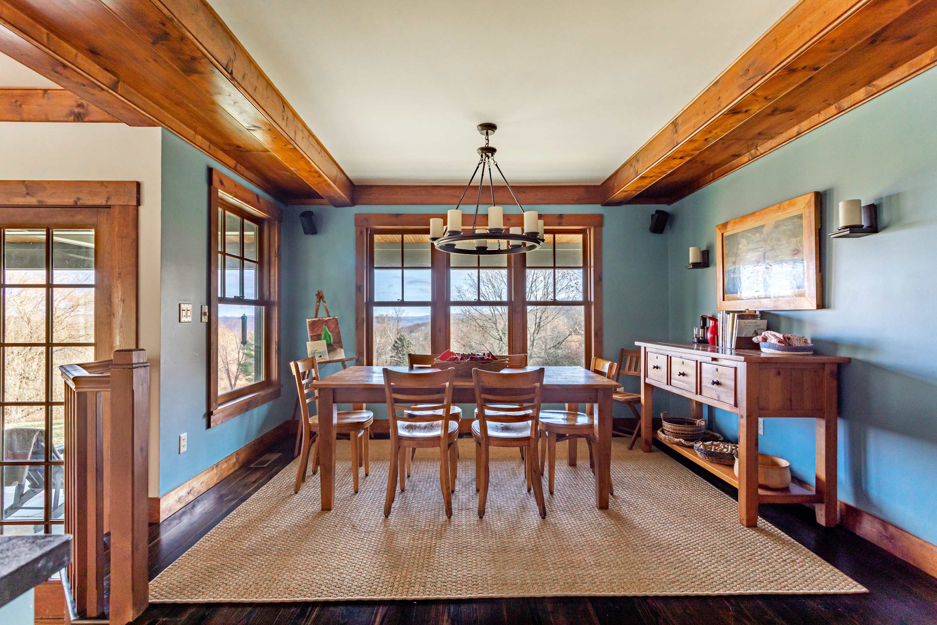 A bright dining room with light blue walls and exposed wooden beams features a large wooden dining table with six chairs, set on a natural fiber rug, with multiple windows offering outdoor views.