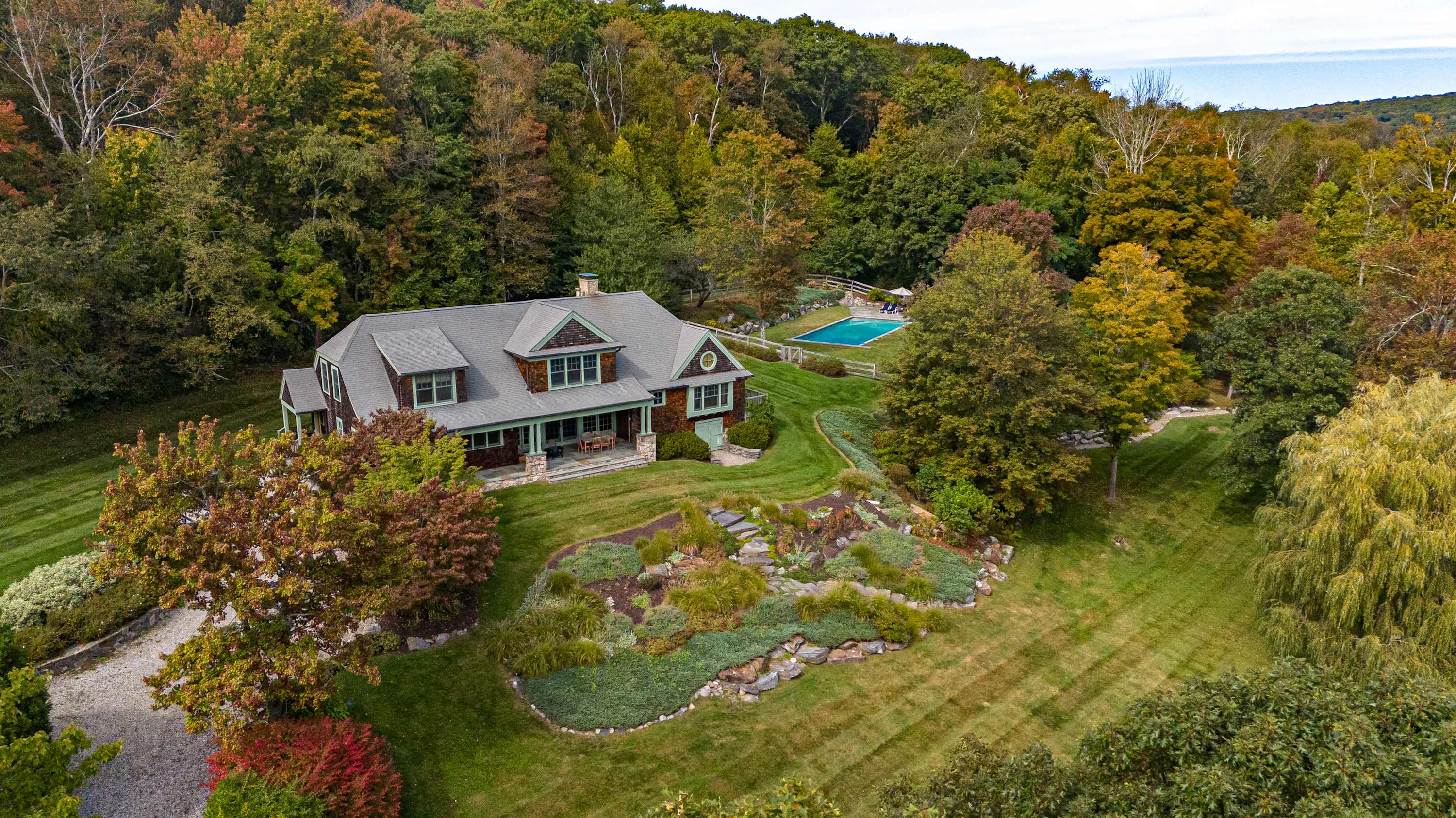 An impressive aerial view showcases the expansive property, featuring the main house with its distinctive green roof, a sparkling blue swimming pool, and meticulously landscaped gardens, all set within a vibrant autumn forest.