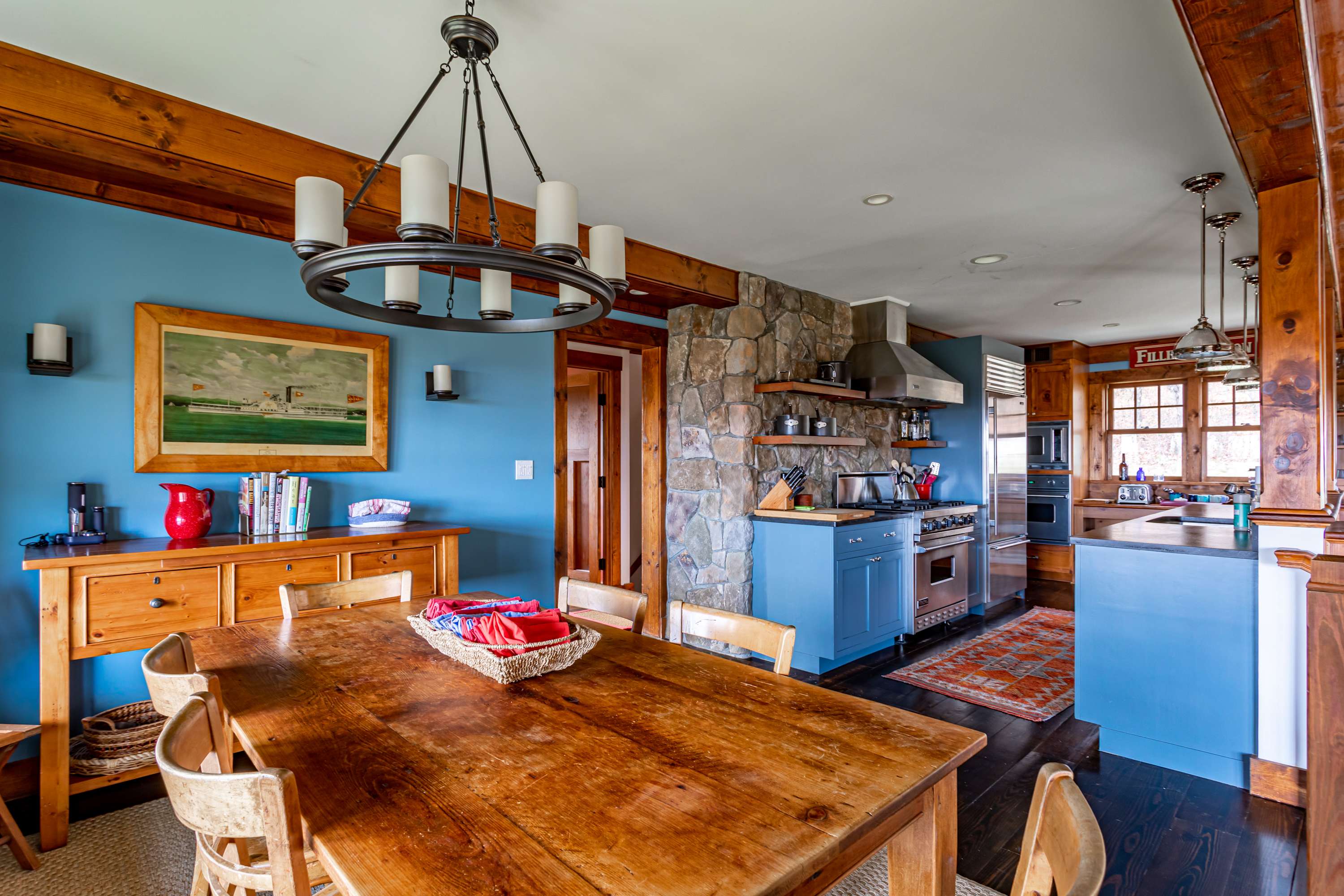 This view of the dining room highlights its connection to the kitchen, featuring a wooden dining table, light blue walls, and exposed wooden beams, with a stone-clad wall visible in the background.