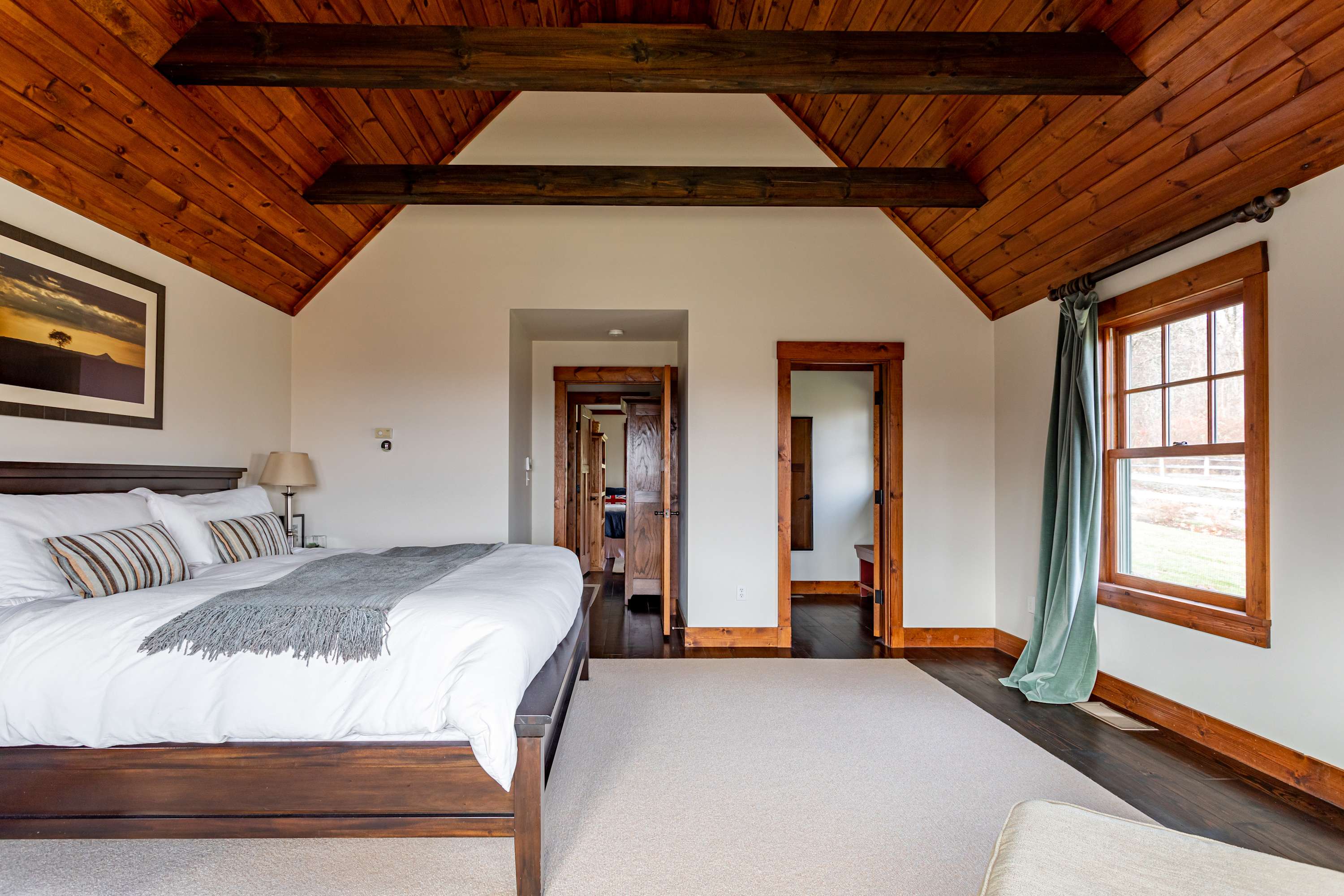 A bright bedroom features a large bed with white bedding, dark hardwood floors, a vaulted wooden ceiling with exposed beams, and a window offering outdoor views.