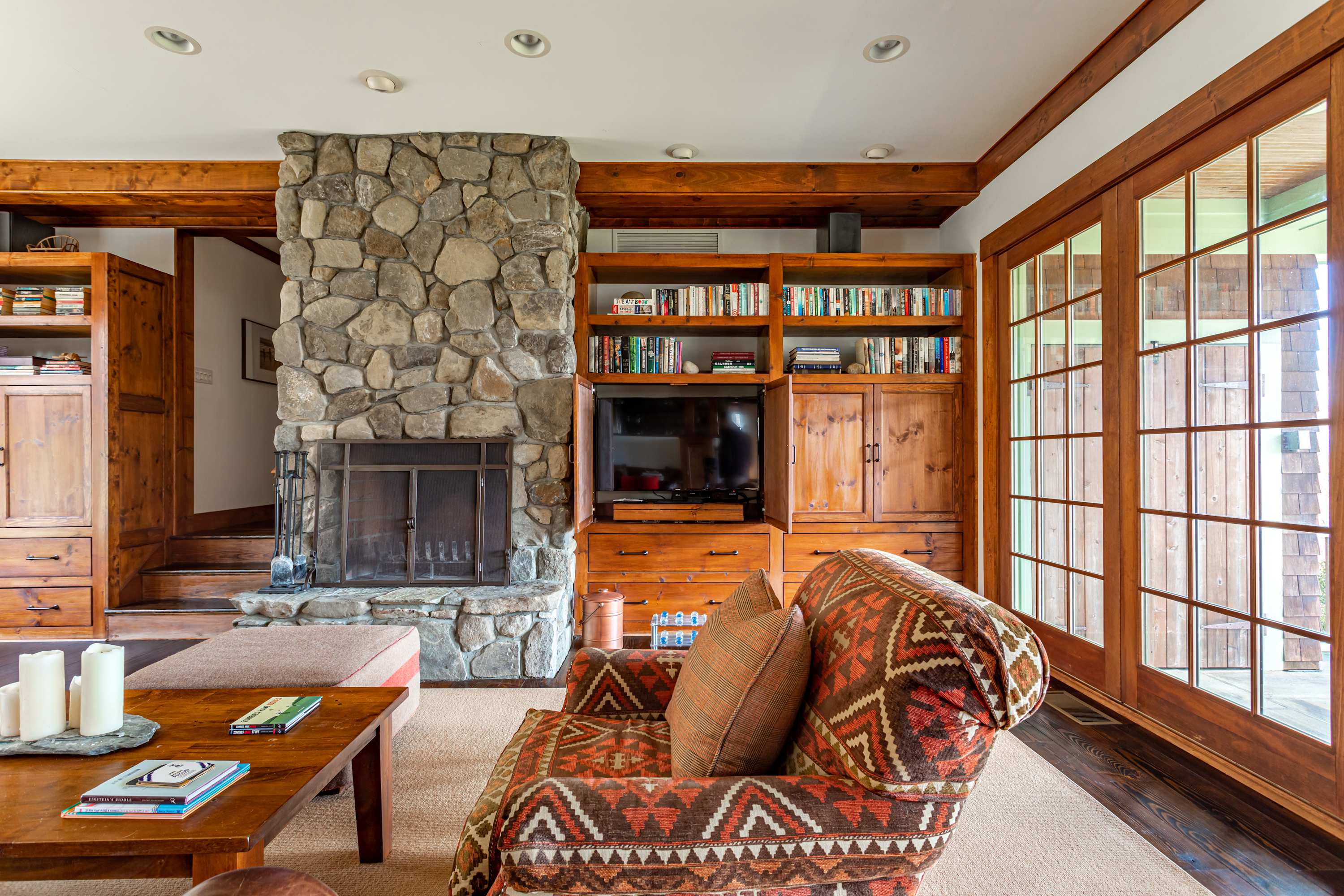 A rustic living room centers around a large stone fireplace, flanked by built-in wooden shelving and cabinets, with a patterned armchair and large glass doors leading outside.
