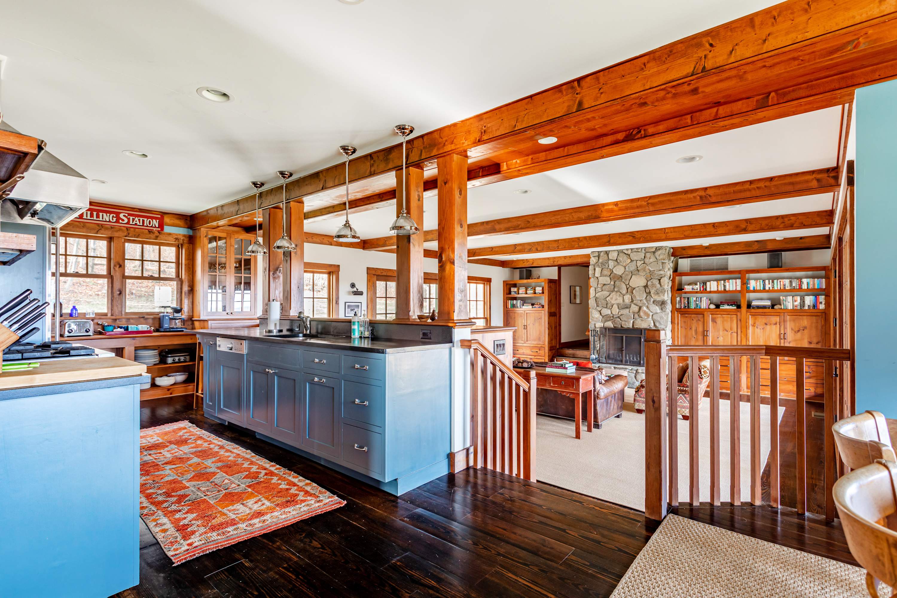 The kitchen, with its blue cabinetry and dark countertops, opens up to a lower-level living area, connected by a wooden staircase and framed by exposed wooden beams and posts.