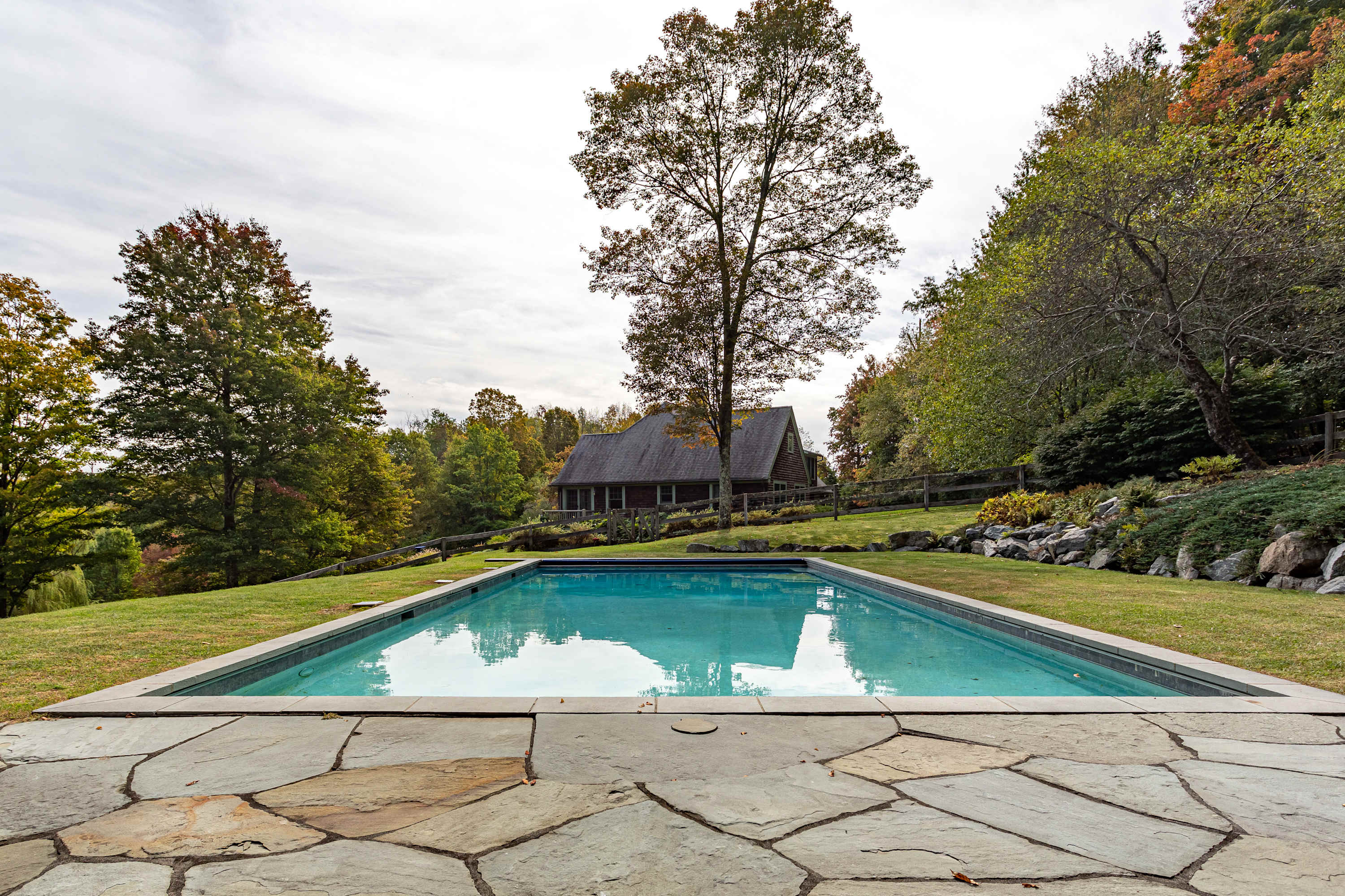 A rectangular swimming pool with a flagstone surround, looking towards the shingle-sided house nestled among trees in the distance under a cloudy sky.