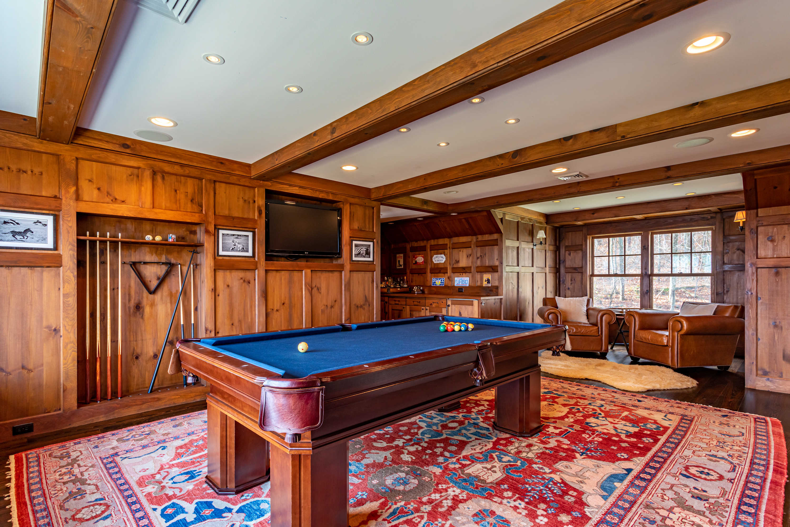 Spacious game room with rich wood paneling and exposed beams, featuring a blue-felt pool table in the center. A wall-mounted TV, leather armchairs, and a vibrant patterned rug complete the inviting space.