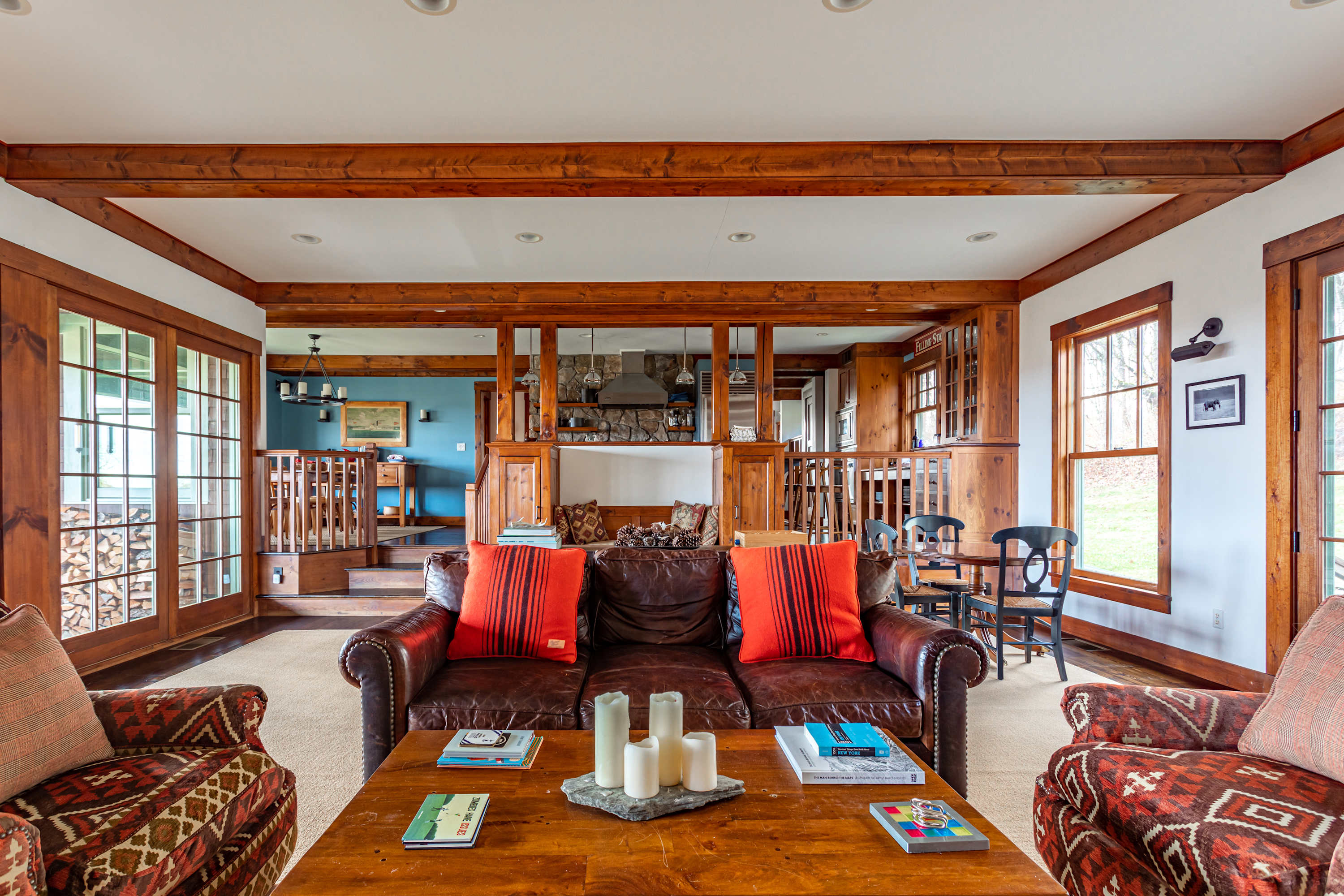 A spacious living room features a brown leather sofa with red throw pillows, two patterned armchairs, a wooden coffee table, and exposed wooden beams, with an open view to the kitchen and dining areas.