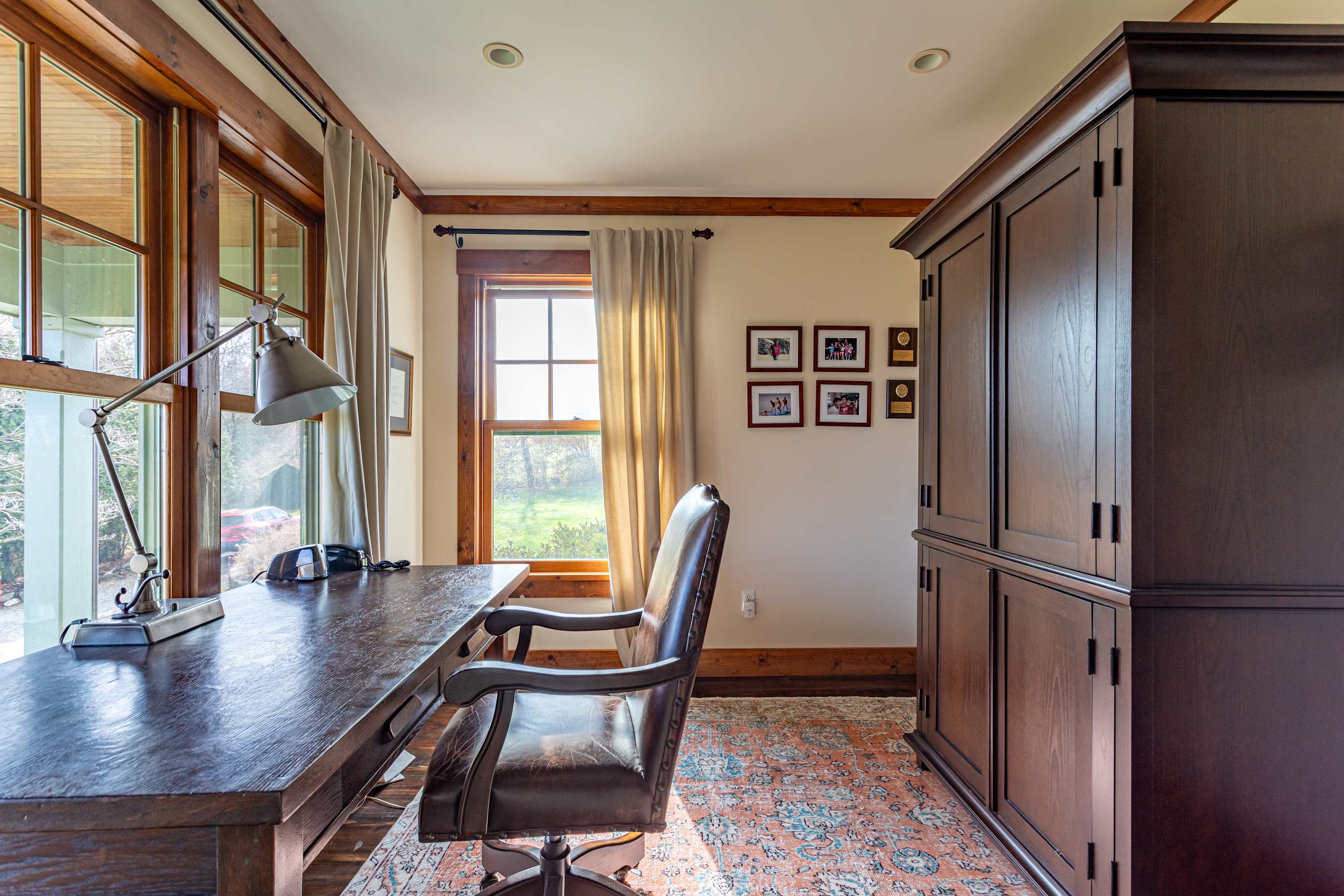 A cozy home office features a large wooden desk with a leather office chair, multiple windows providing natural light and outdoor views, and a tall wooden cabinet.