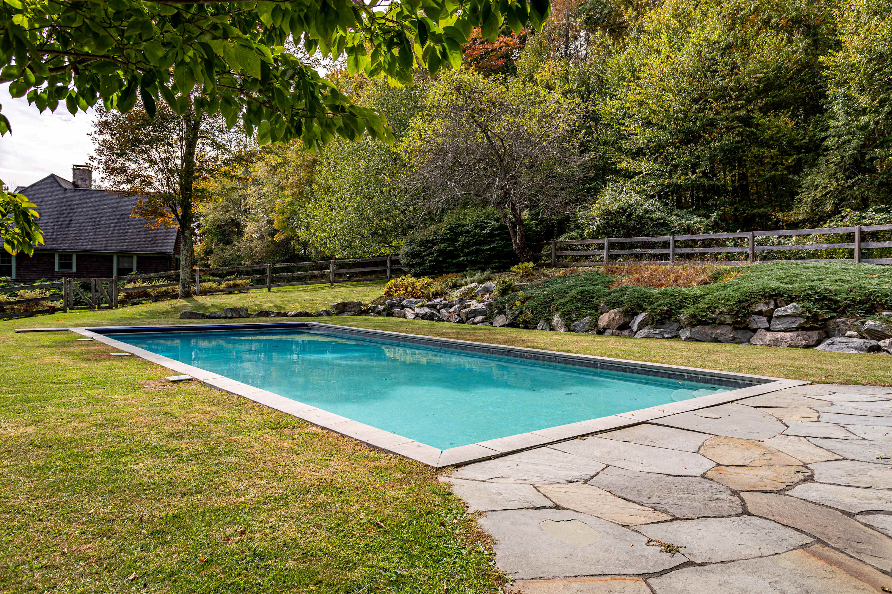 A long rectangular swimming pool set in a grassy lawn, bordered by a flagstone path, with a stone retaining wall and a wooden fence leading to a wooded area, and the house visible in the background.