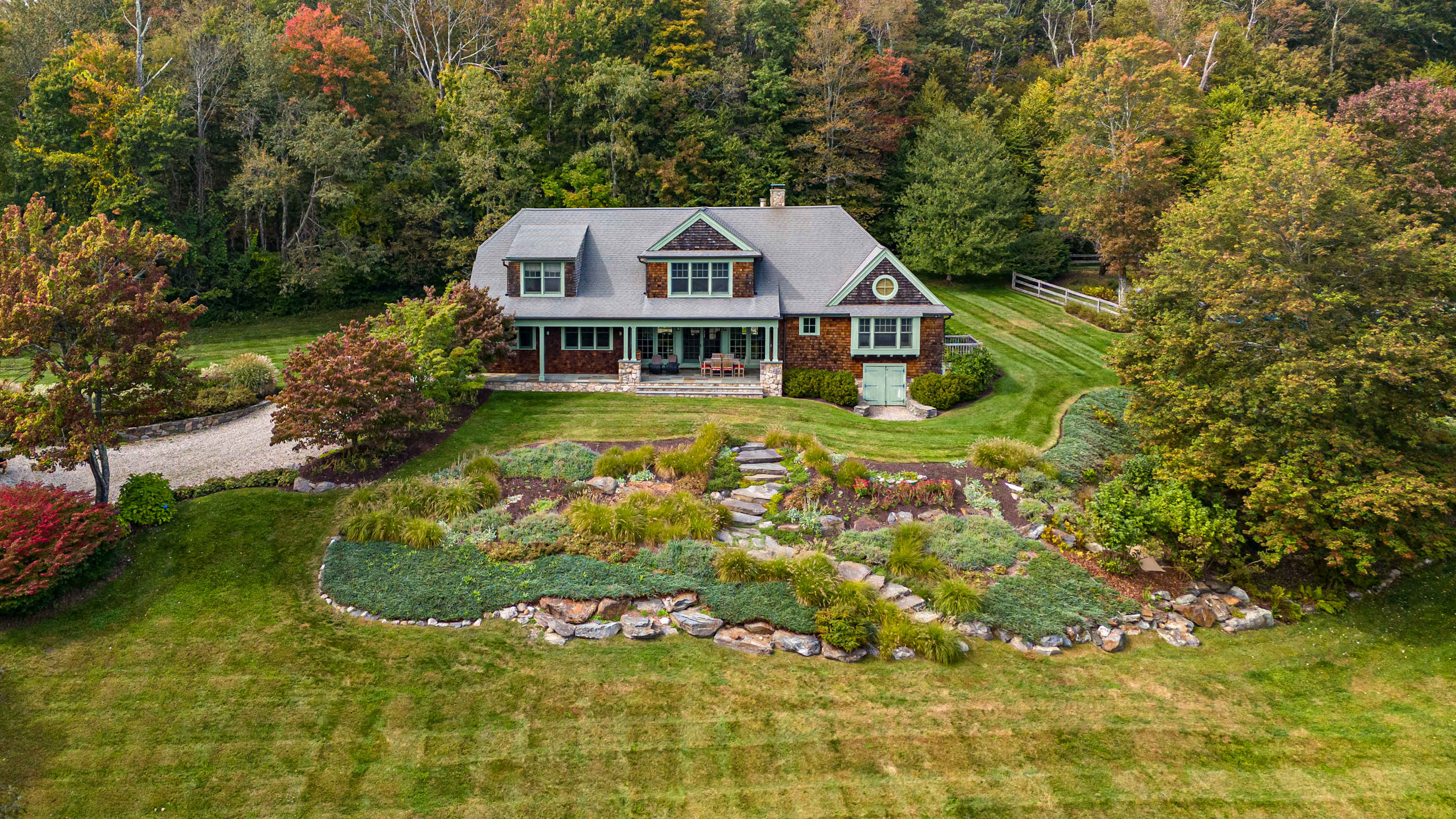 An aerial view showcases the shingle-sided house with green trim, nestled amidst a meticulously landscaped terraced garden and a gravel driveway, surrounded by lush green and autumn-colored trees.