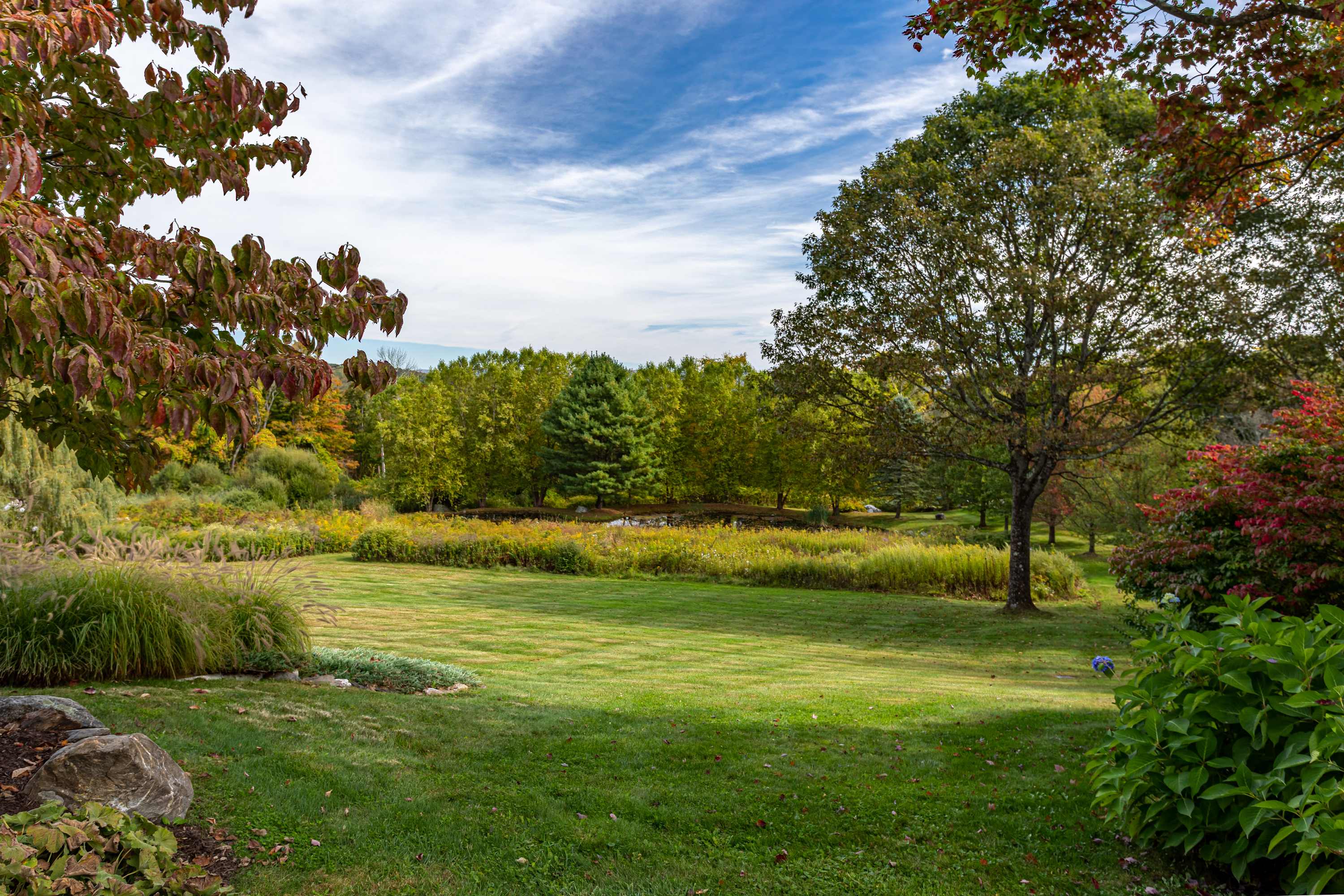 An expansive green lawn stretches towards a distant pond and dense forest, with a variety of trees displaying early autumn colors under a bright blue sky.