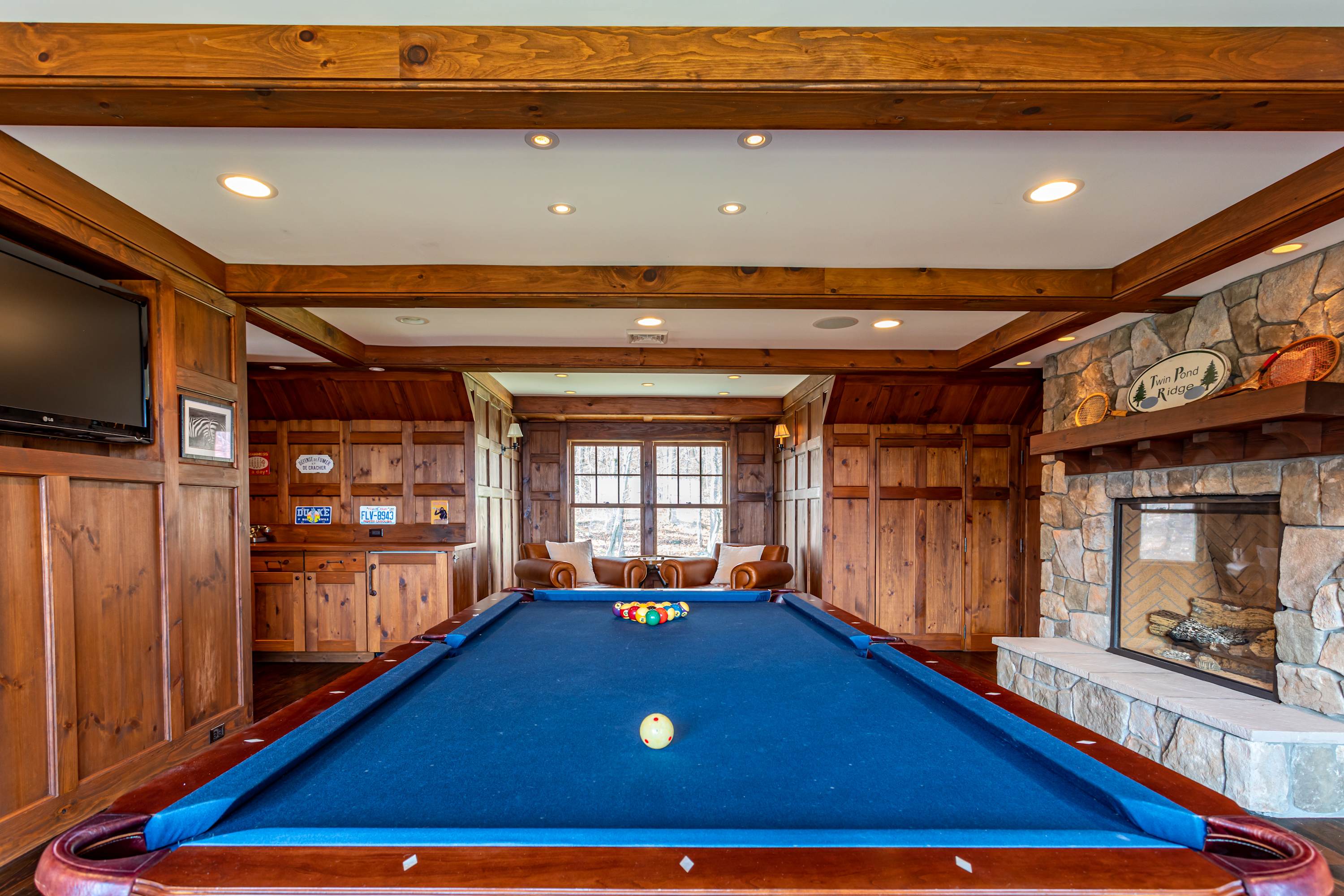 Wide view of the game room showcasing a blue-felt pool table, a built-in wooden bar area, and a grand stone fireplace. Large windows at the far end offer views of the outdoors, with leather armchairs arranged nearby.