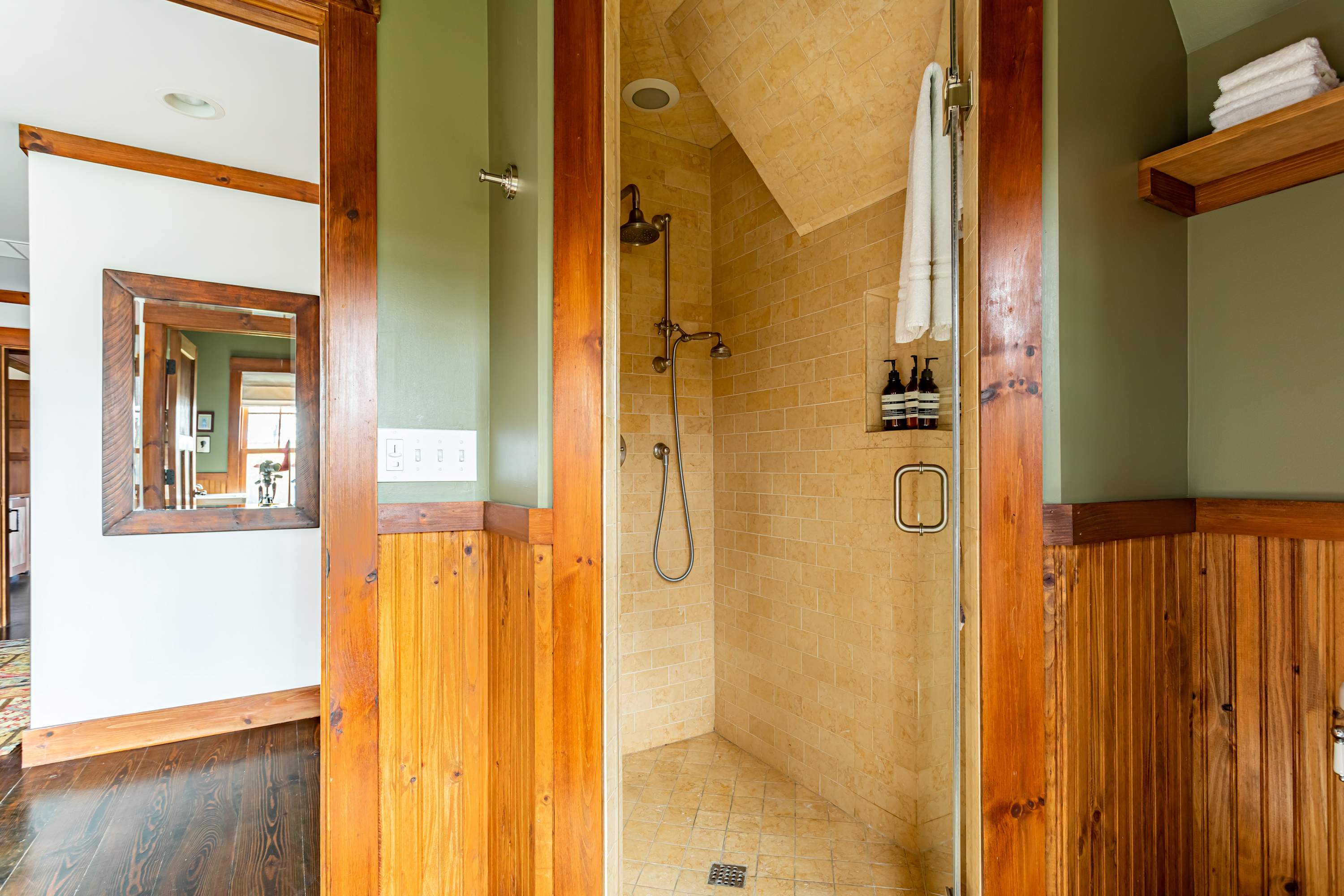 A walk-in shower with light beige tiled walls and floor, featuring a rain showerhead and a handheld shower, framed by rustic wooden trim and olive green walls, adjacent to a wooden wainscoted area.