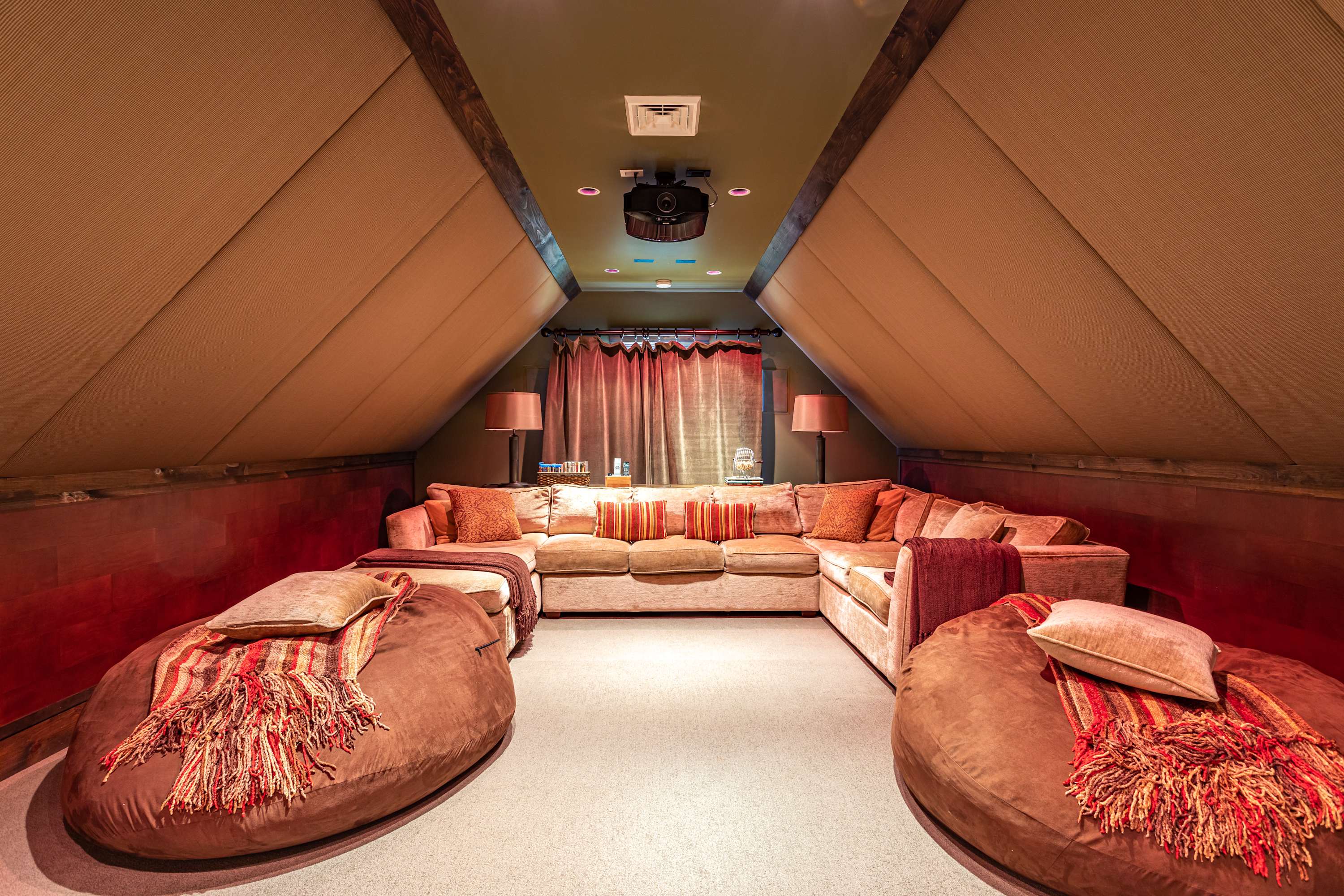 A dedicated media room features sloped ceilings with acoustic paneling and warm lighting, creating an immersive viewing experience. A large, comfortable sectional sofa and two oversized bean bags provide ample seating for movie nights.