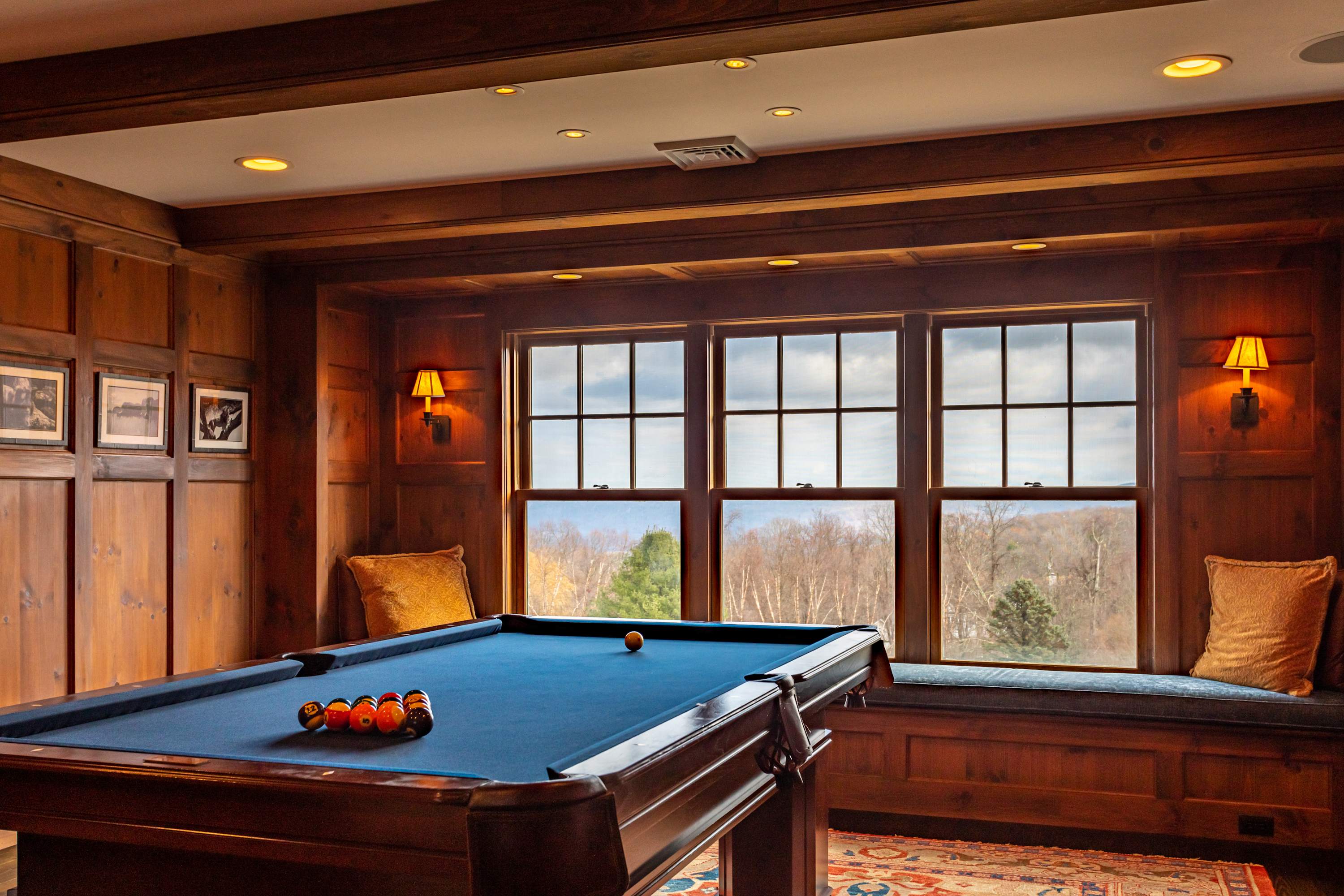A sophisticated game room features a dark wood pool table with a vibrant blue felt surface, set against rich wood-paneled walls. Large windows with a built-in window seat offer expansive views of the distant wooded landscape.