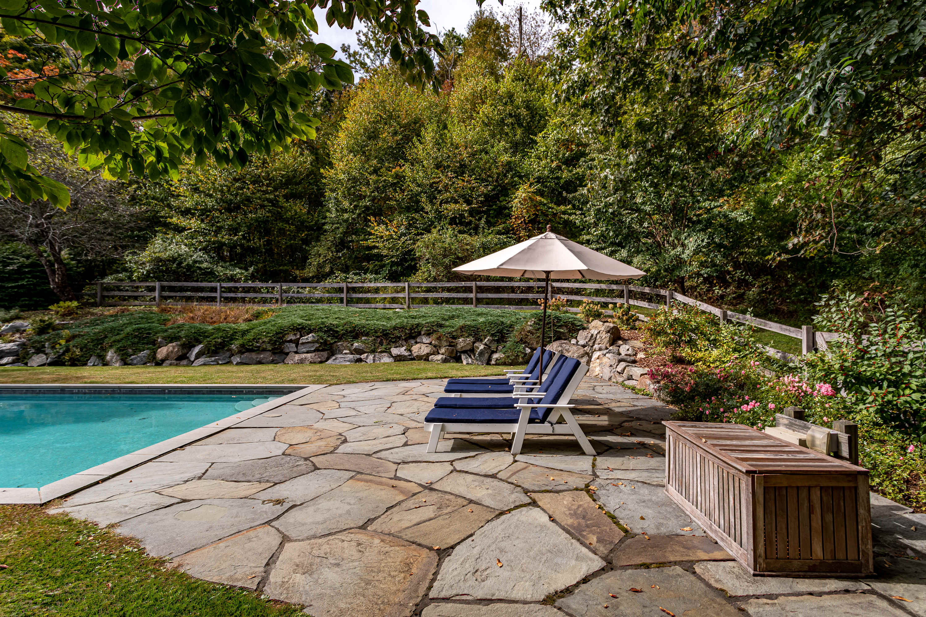 A close-up view of the swimming pool area highlights a rustic flagstone patio furnished with two blue lounge chairs and a cream-colored umbrella, surrounded by lush green landscaping and a wooden fence.