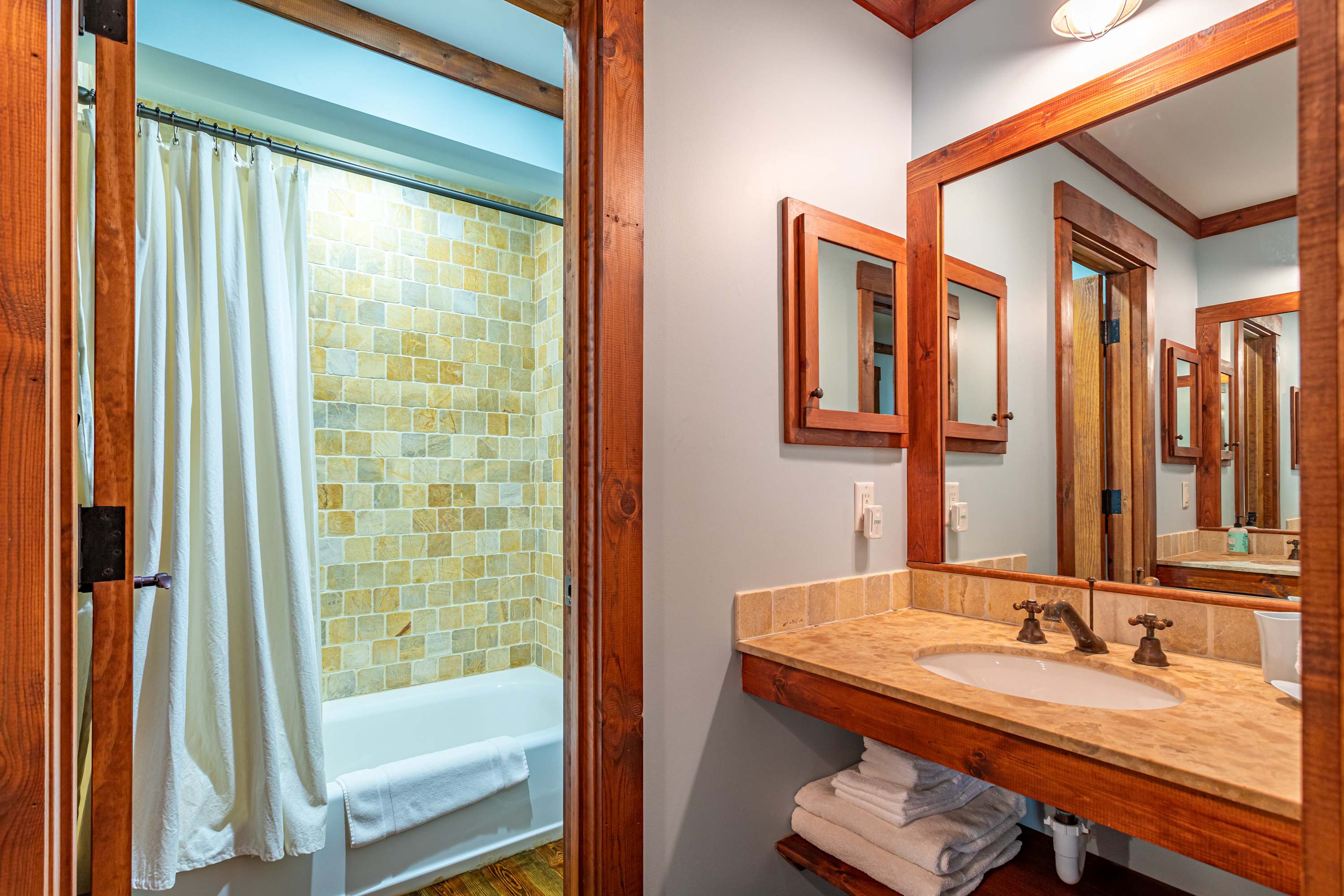 A bathroom with a single vanity sink set in a light-colored countertop with a wooden base, a large mirror, and a view into a shower/tub combo with light square tiled walls and a white shower curtain.