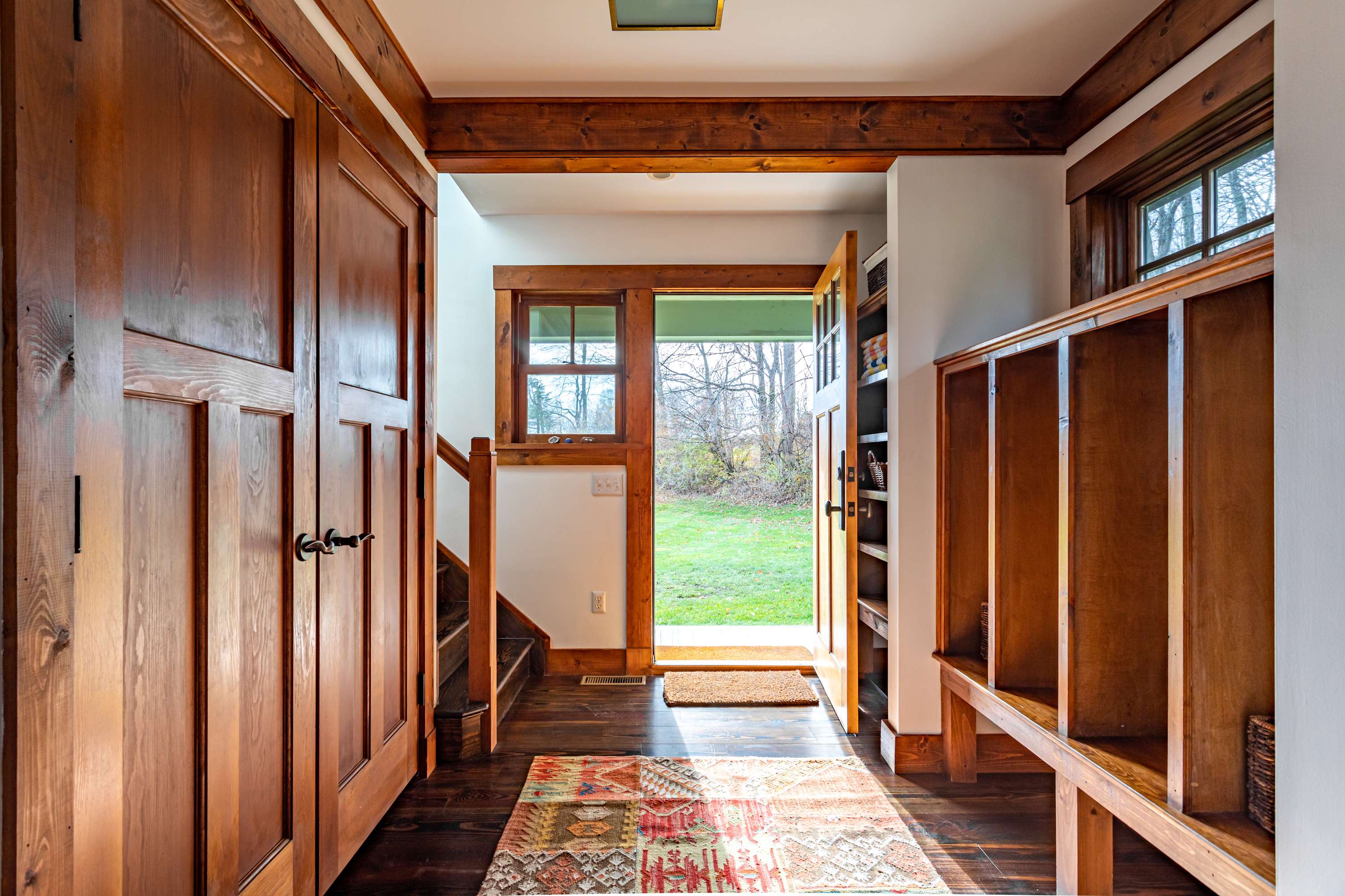 A warm and inviting entryway features rich wooden paneling and built-in storage, with a colorful rug leading to an open door revealing a grassy outdoor area.