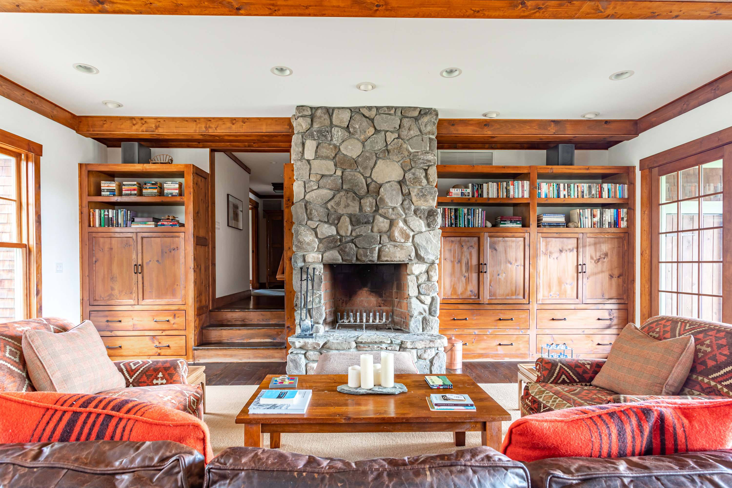A grand living room is anchored by a floor-to-ceiling stone fireplace, flanked by custom built-in wooden bookshelves and cabinets. Two comfortable leather sofas with patterned pillows face each other, creating a cozy and inviting seating arrangement.