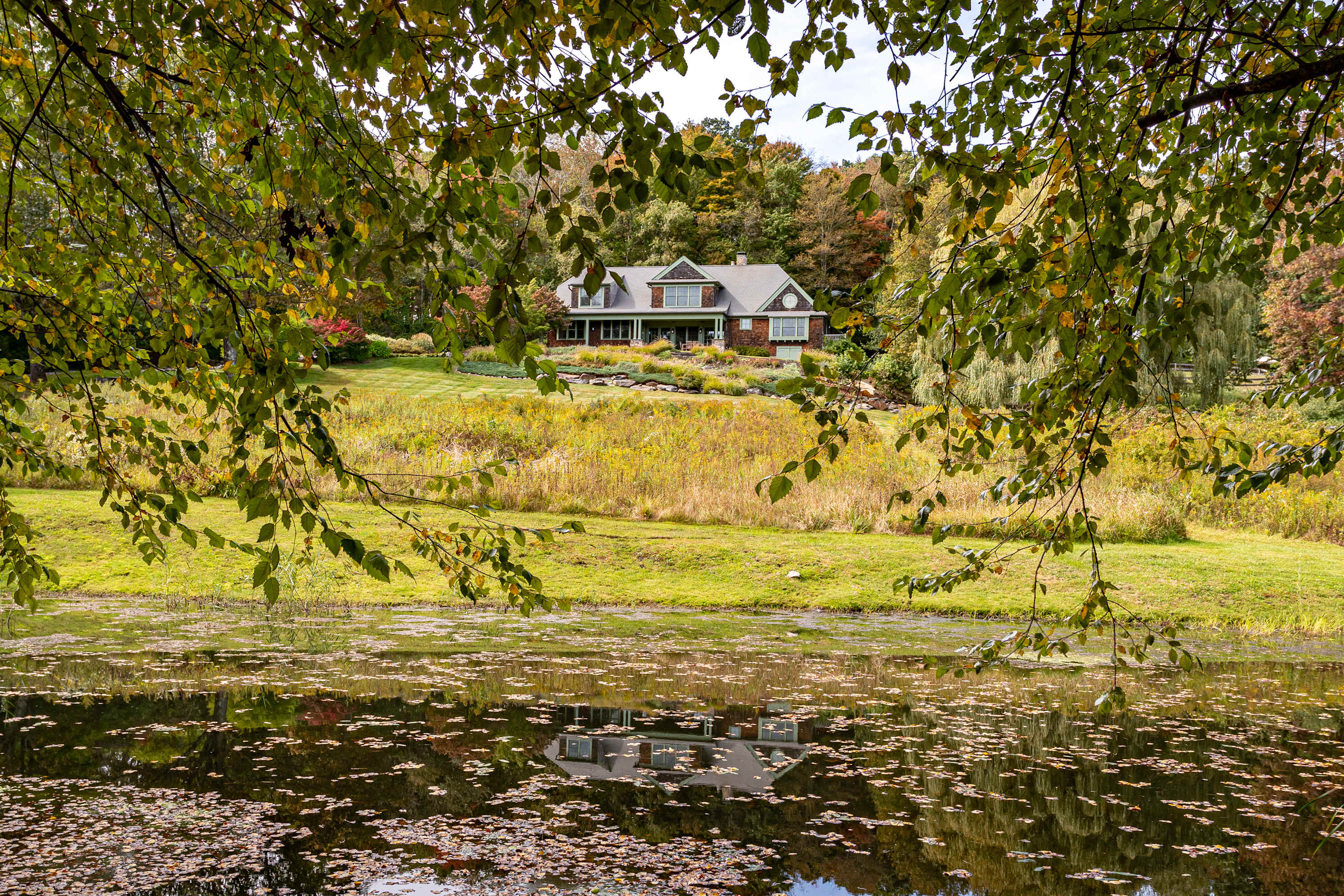 The house is reflected in the calm waters of a pond, framed by overhanging tree branches, with a field of tall golden grasses and autumn trees in the background.