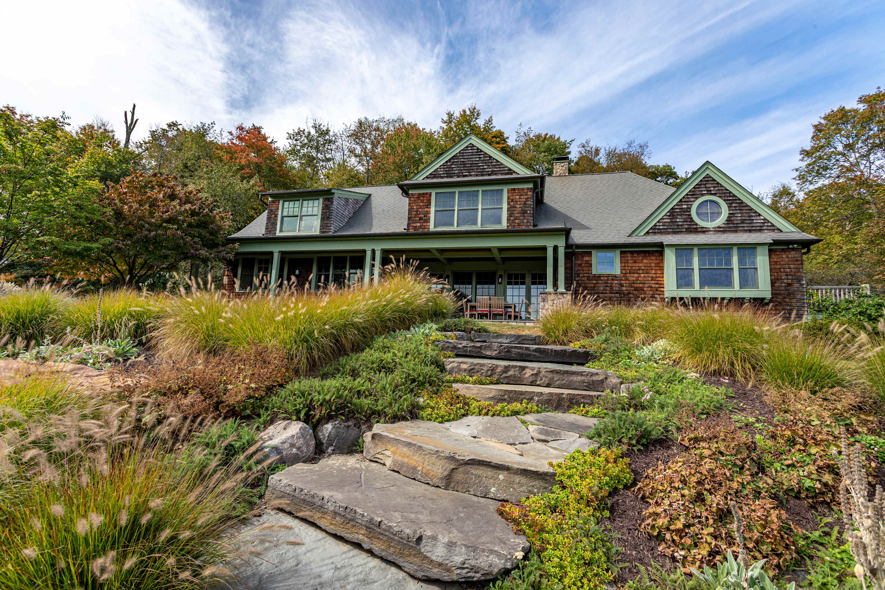 A charming shingle-sided house with green trim and a stone pathway leading up a landscaped hill, surrounded by lush trees and a partly cloudy sky.