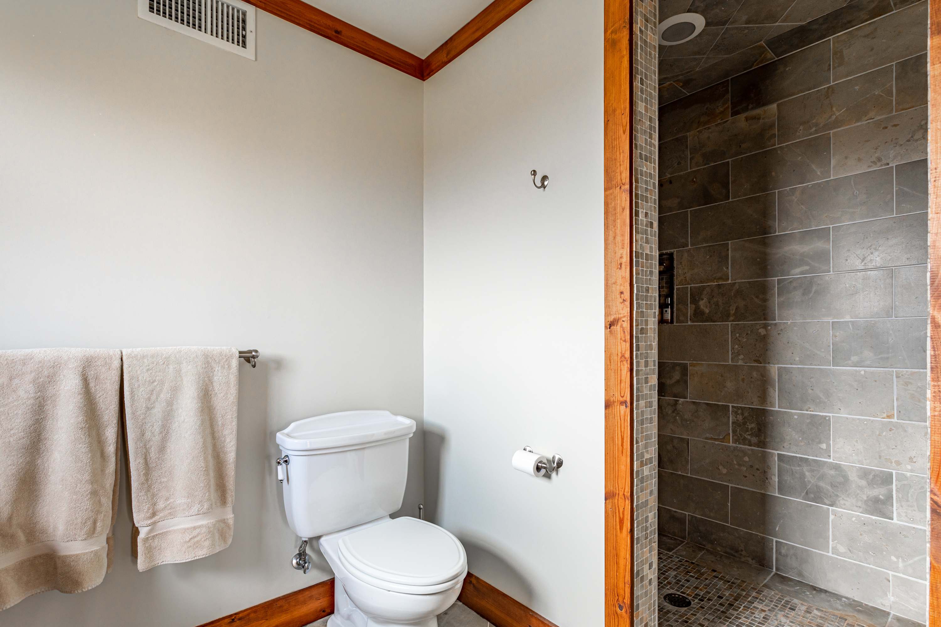 A modern bathroom featuring a white toilet, light beige walls, and a walk-in shower with dark gray rectangular tiles and a mosaic-tiled floor, framed by rustic wooden trim.