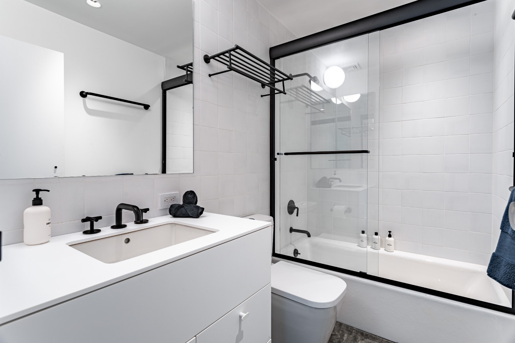 A different angle of the modern bathroom, emphasizing the white subway tiled shower/tub combo with a black-framed glass door. The white vanity with a black faucet and mirror maintains the clean, minimalist aesthetic.