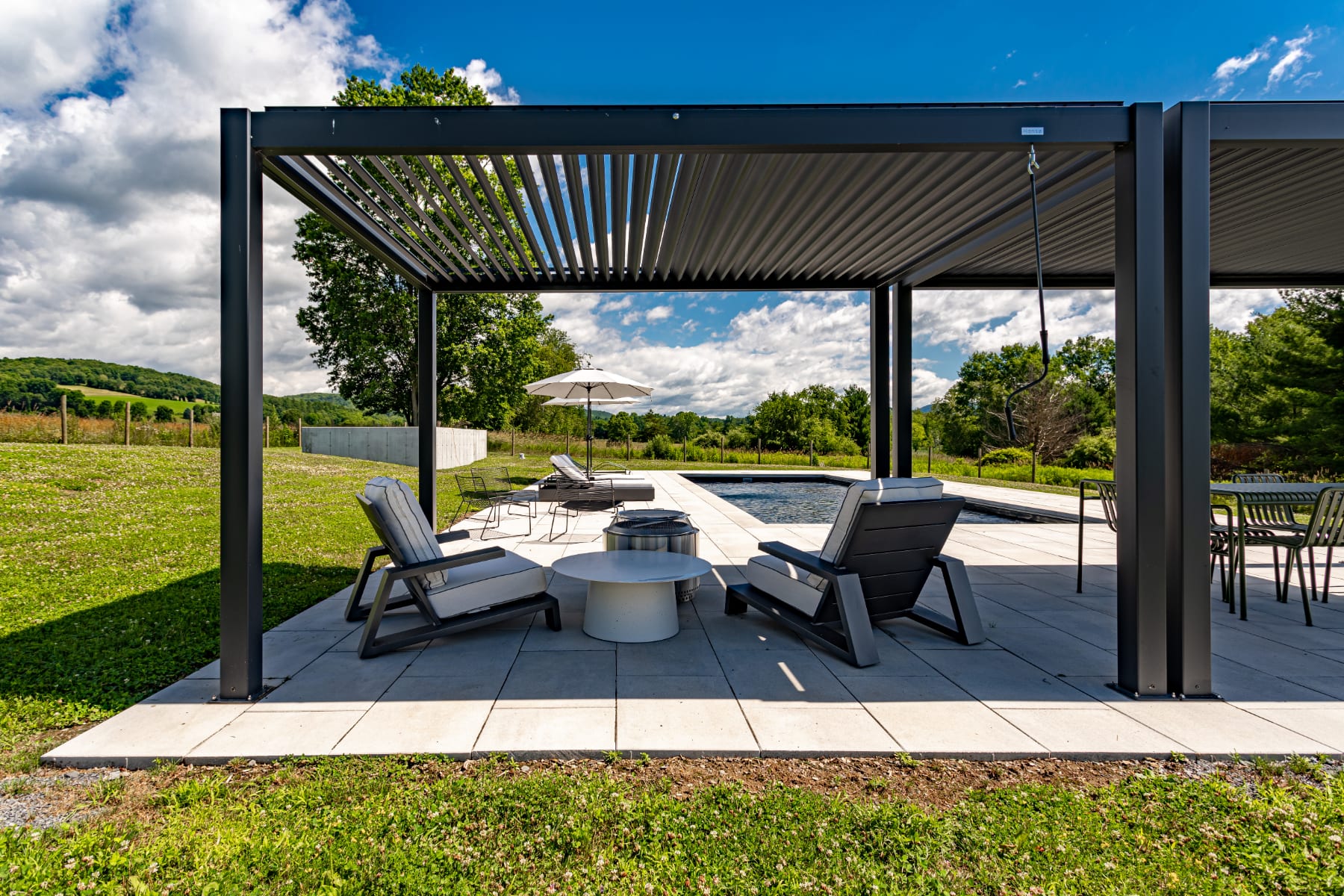 Stylish outdoor lounge area under a modern black slatted pergola, featuring two grey lounge chairs and a white round coffee table. The space offers views of the swimming pool, green fields, and distant mountains.