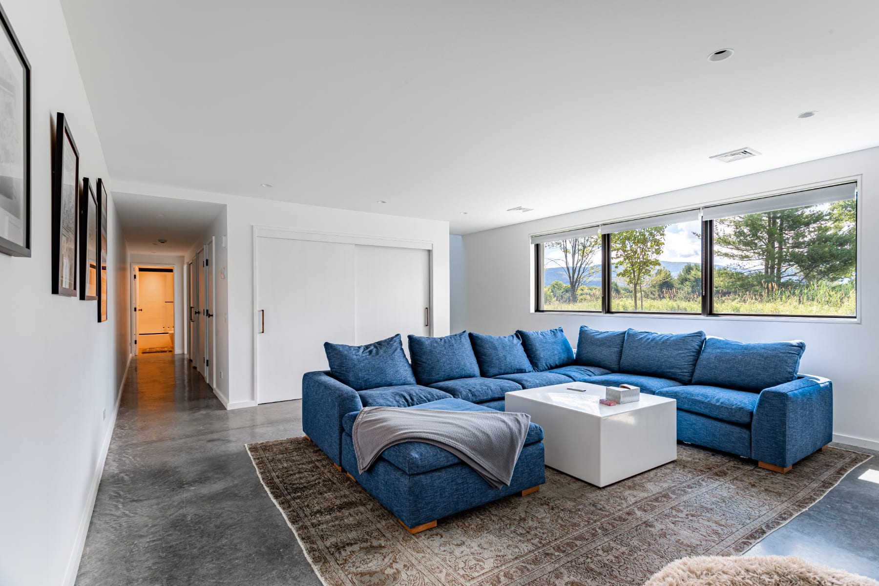 Spacious living room featuring a large blue sectional sofa, a minimalist white coffee table, and wide windows offering views of the outdoors, set on a patterned rug over a concrete floor.
