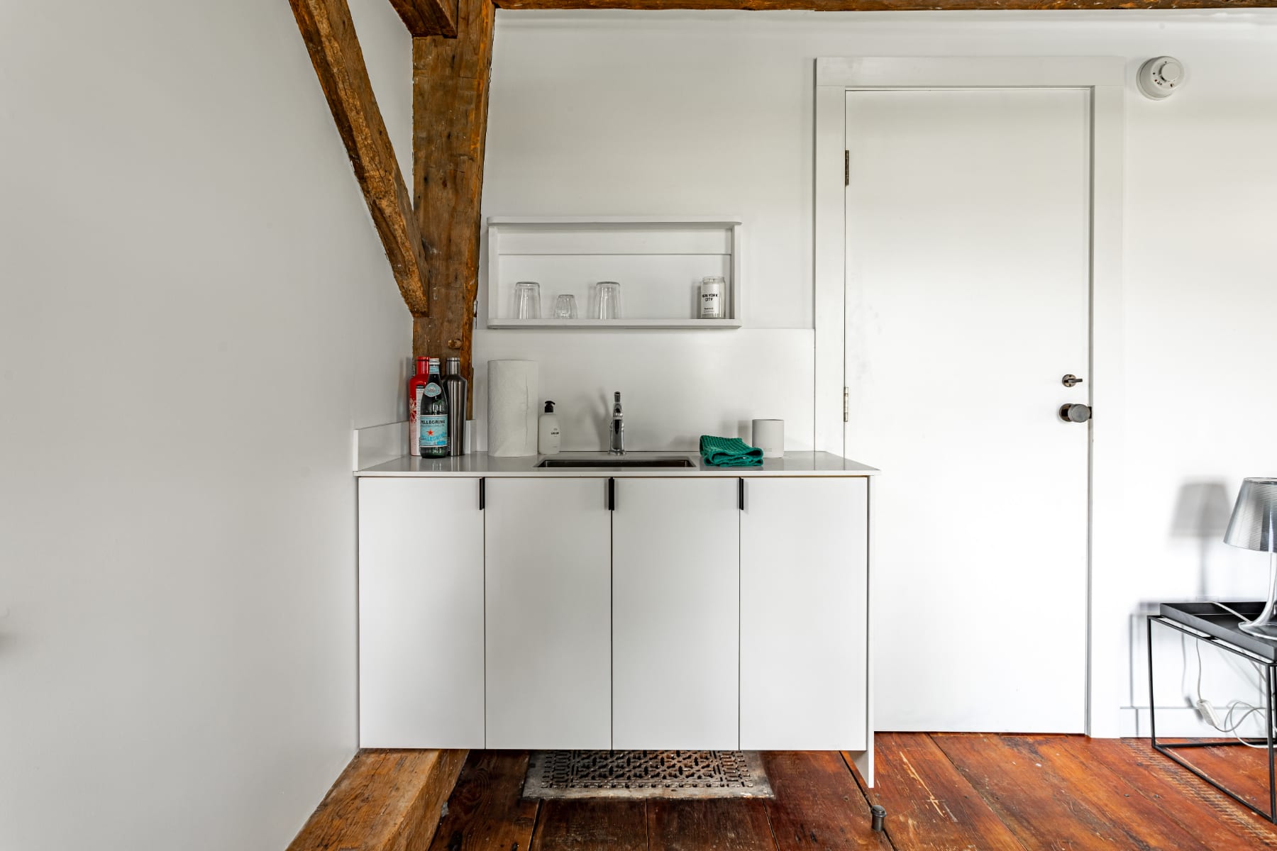 Compact white kitchenette with a sink, minimalist cabinetry, and a small shelf, set against a white wall with rustic exposed wooden beams overhead.