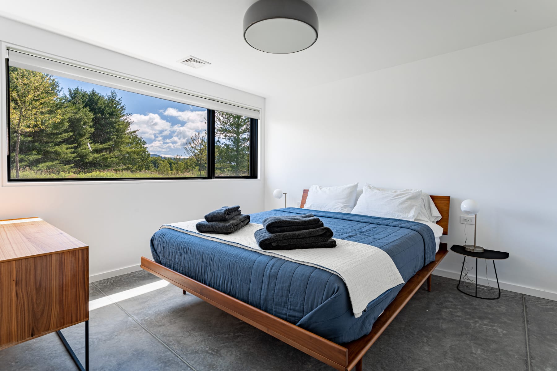 Bright and airy bedroom featuring a wooden platform bed with dark blue and white bedding, set against a backdrop of white walls and polished concrete floors. A wide horizontal window offers a panoramic view of trees and mountains.