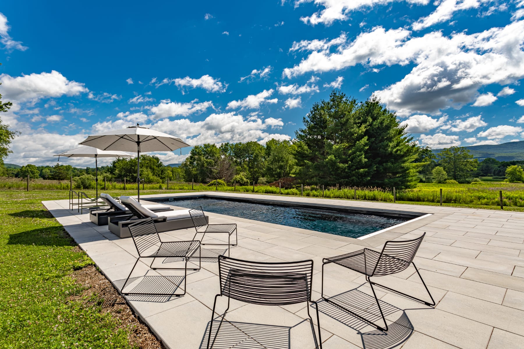 Inviting rectangular swimming pool set on a light-colored stone patio, surrounded by comfortable lounge chairs and white umbrellas. The expansive green lawn and distant mountains create a serene, sunny outdoor oasis.