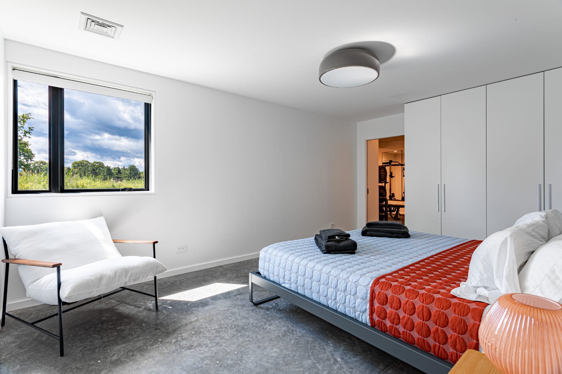 Another view of the modern bedroom, showcasing a large window with a view of lush greenery. A white armchair with a black frame sits by the window, and sleek white built-in wardrobes are visible near the doorway.