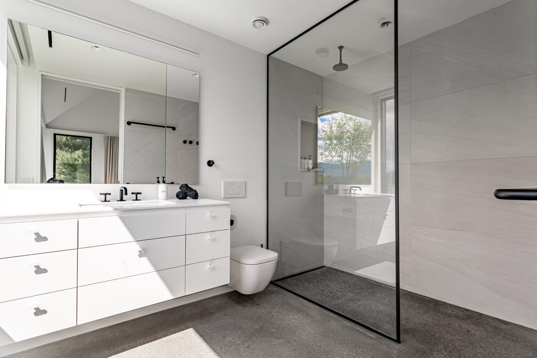 Contemporary bathroom with a floating white vanity, a toilet, and a spacious walk-in shower with a glass enclosure, all set on a concrete floor with ample natural light.
