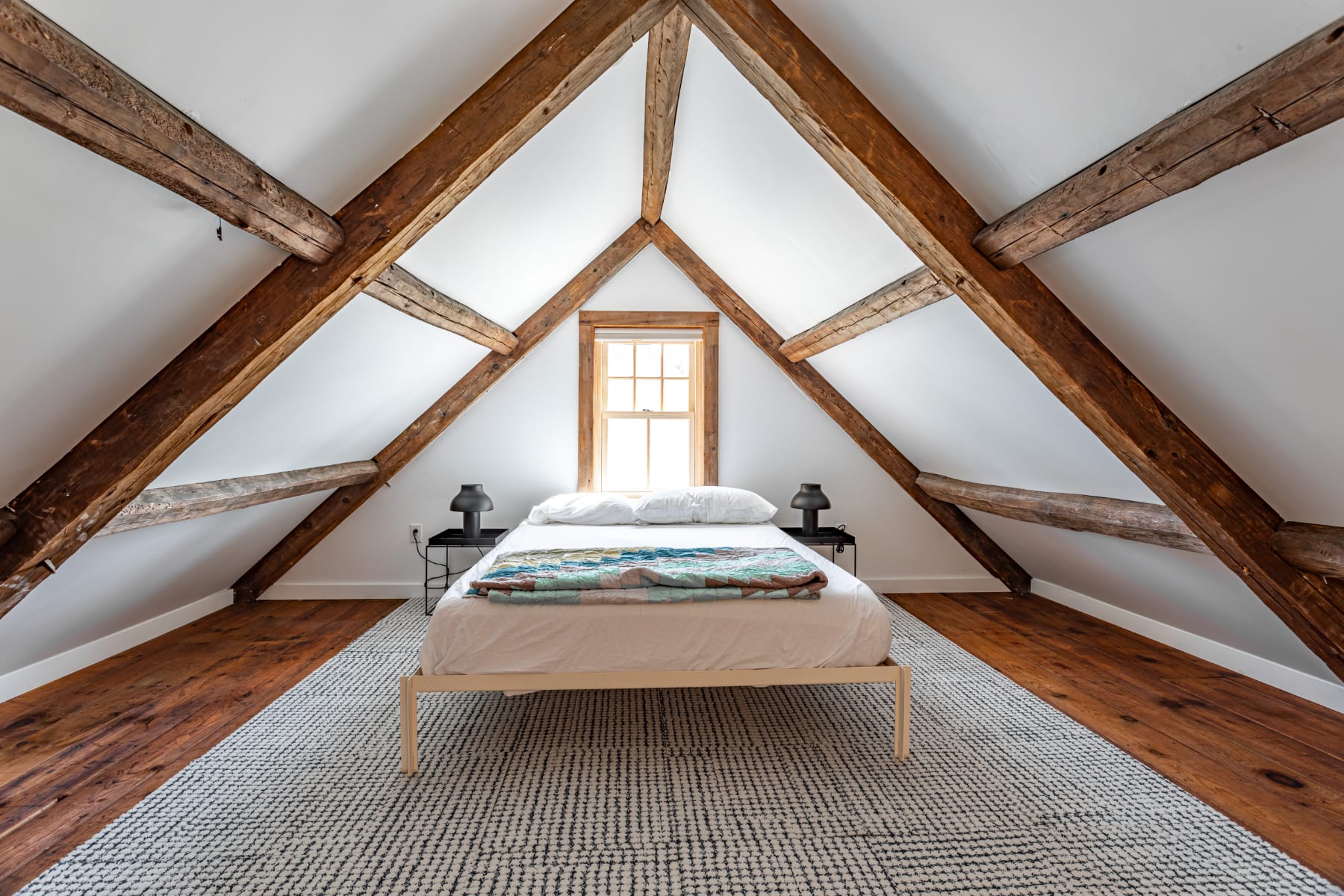 Serene bedroom with a minimalist bed, white bedding, and a colorful quilt, featuring a dramatic vaulted ceiling with exposed wooden beams, white walls, and warm hardwood floors.