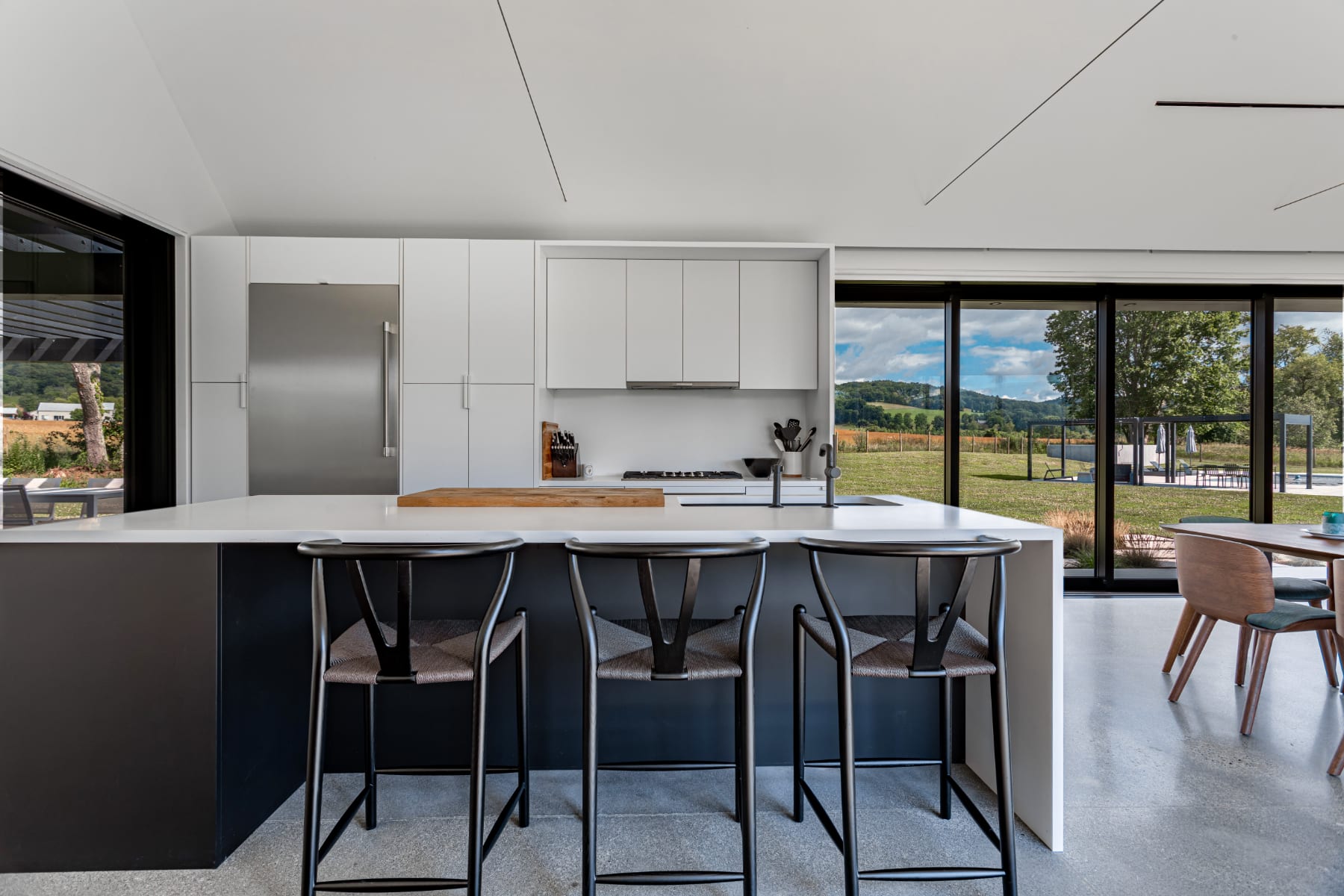 Contemporary kitchen island with a white countertop and dark base, accompanied by three black bar stools, offering a view of the gas range and expansive green landscape through large sliding glass doors.