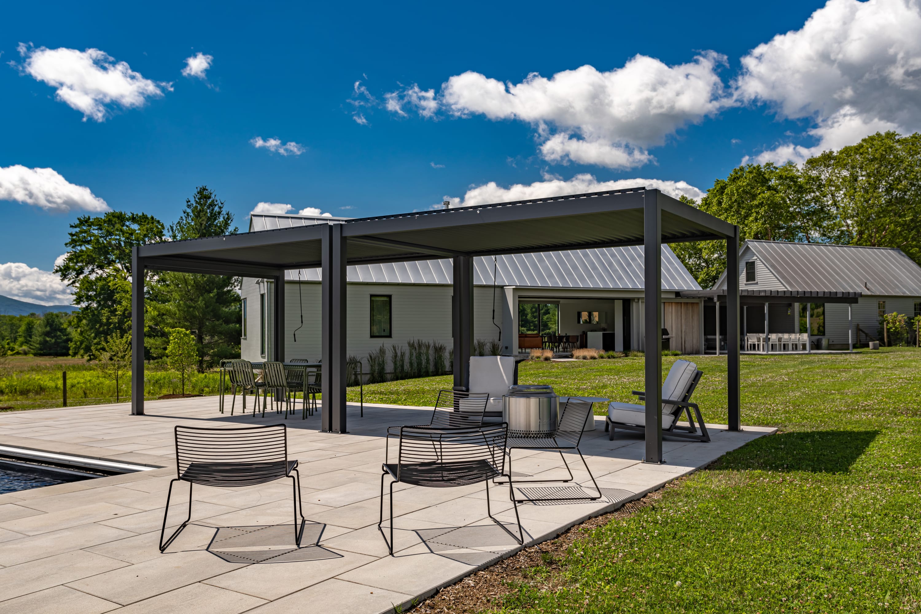 Panoramic view of the property's modern exterior, showcasing a large black pergola covering outdoor dining and lounge areas on a stone patio next to the swimming pool. The house is surrounded by a manicured green lawn and trees.