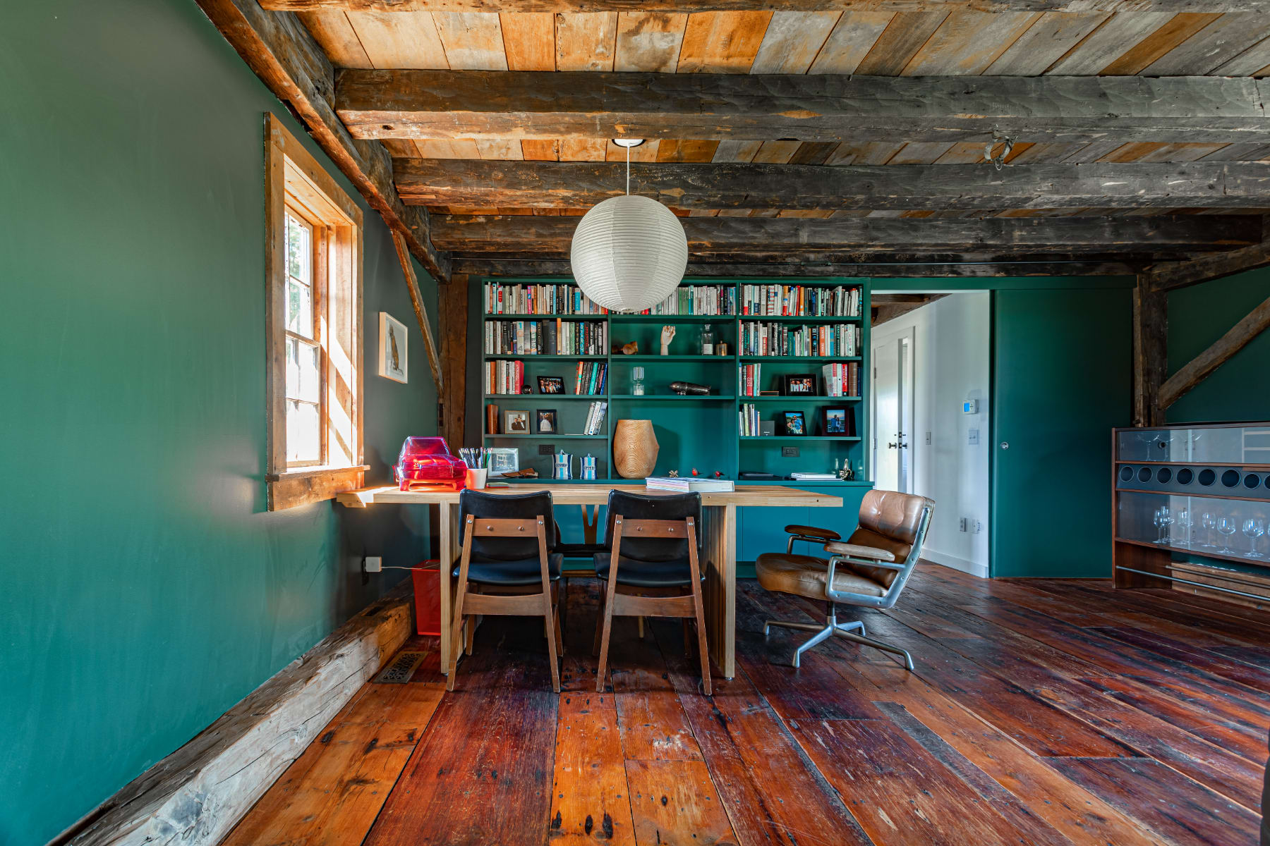 Spacious rustic office with dark green walls, a long wooden desk, and a comfortable leather armchair, surrounded by exposed wooden beams, rich hardwood floors, and a large built-in bookshelf.