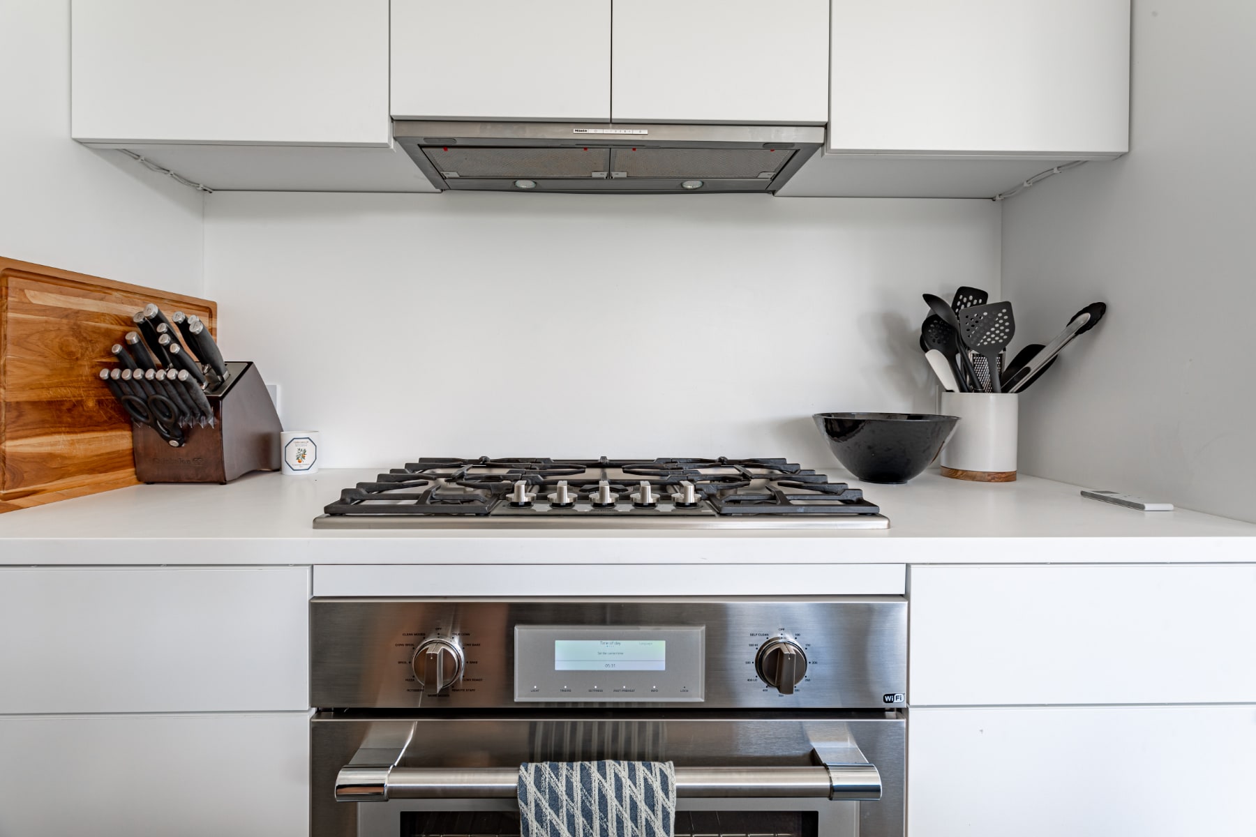 Close-up of a modern kitchen's cooking station, showcasing a stainless steel gas cooktop with six burners, a built-in stainless steel oven, and minimalist white cabinetry above and below.