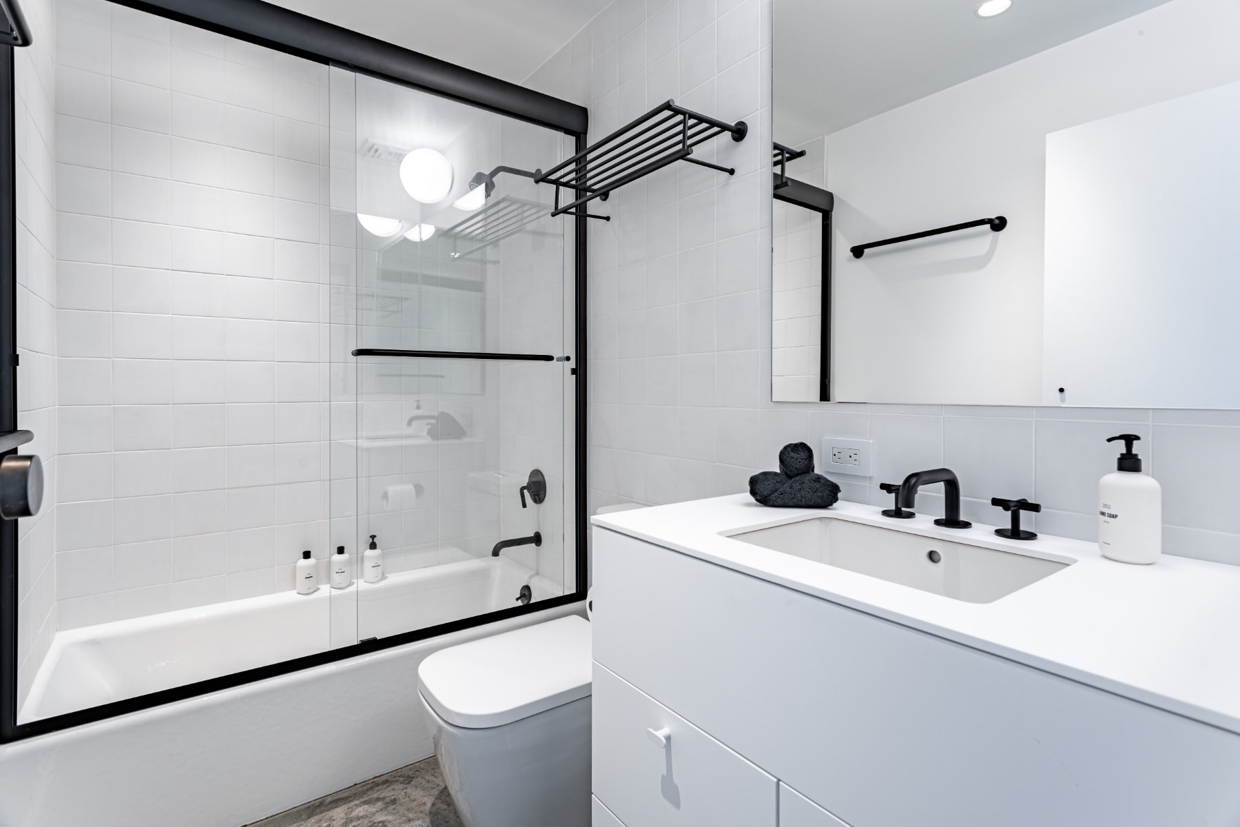 Contemporary bathroom with white subway tile walls, a white vanity with a rectangular sink and black faucet, and a bathtub enclosed by a black-framed sliding glass shower door. Black hardware accents the minimalist design.