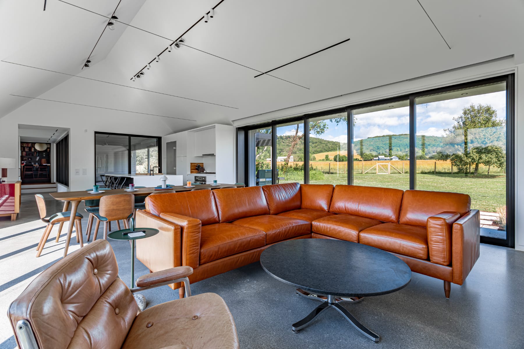 Wide shot of an open-plan living space with a large brown leather sectional, a round coffee table, and an armchair. Expansive floor-to-ceiling windows showcase a panoramic view of rolling green hills and fields under a bright sky.