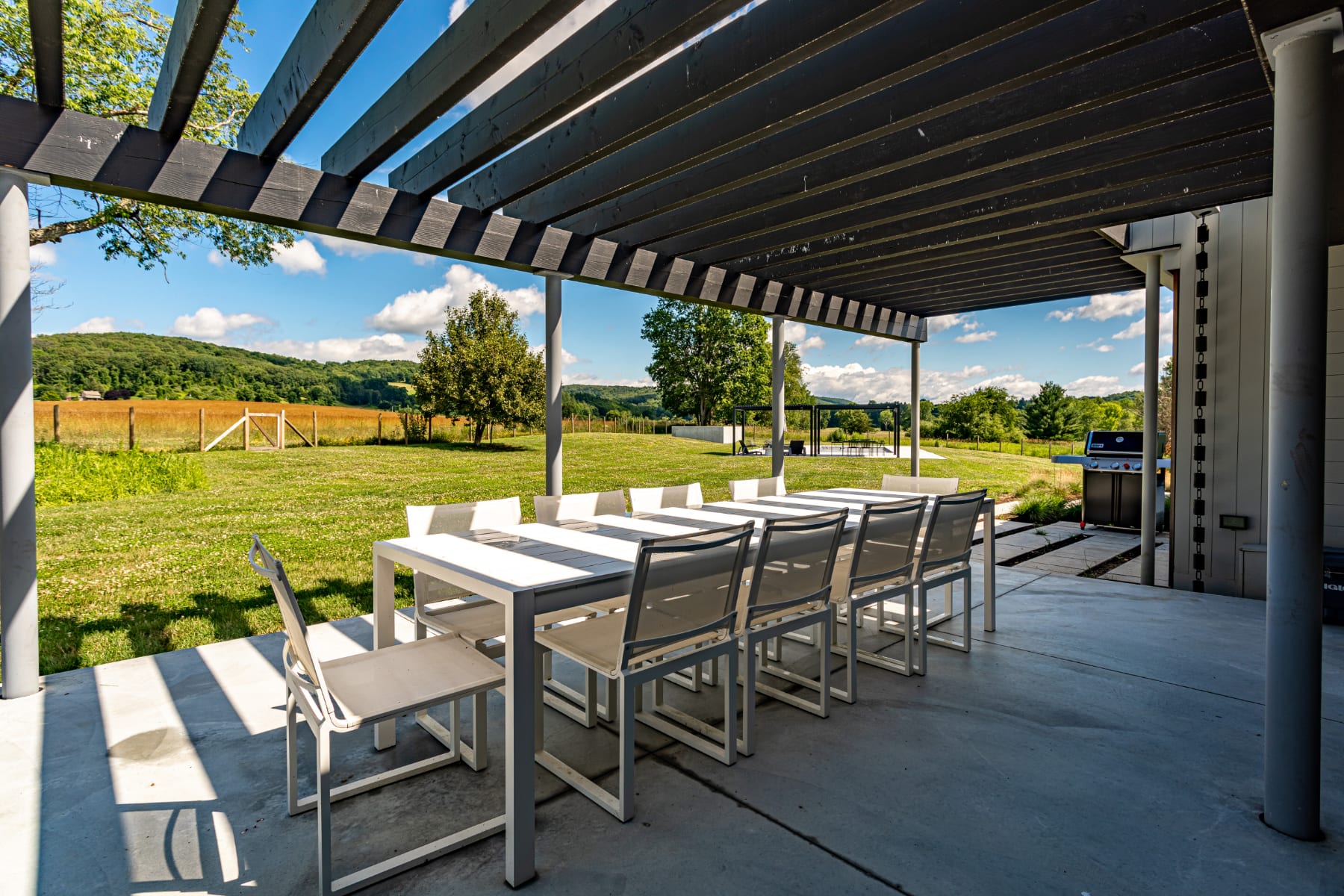 A modern outdoor patio features a long white dining table with ten chairs under a dark wooden pergola, overlooking a vast green lawn and rolling hills under a blue sky. A BBQ grill is visible on the right.