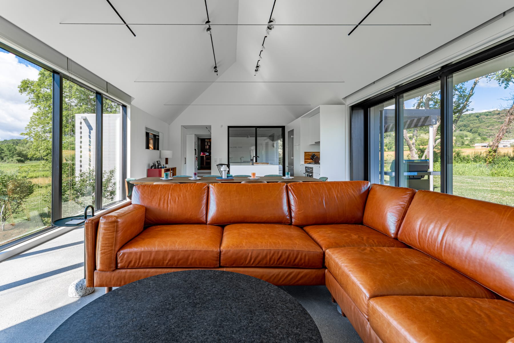 Spacious open-concept living area featuring a large brown leather sectional sofa and a round black coffee table on polished concrete floors. Floor-to-ceiling windows offer views of the surrounding green landscape, with a dining area and kitchen visible in the background.