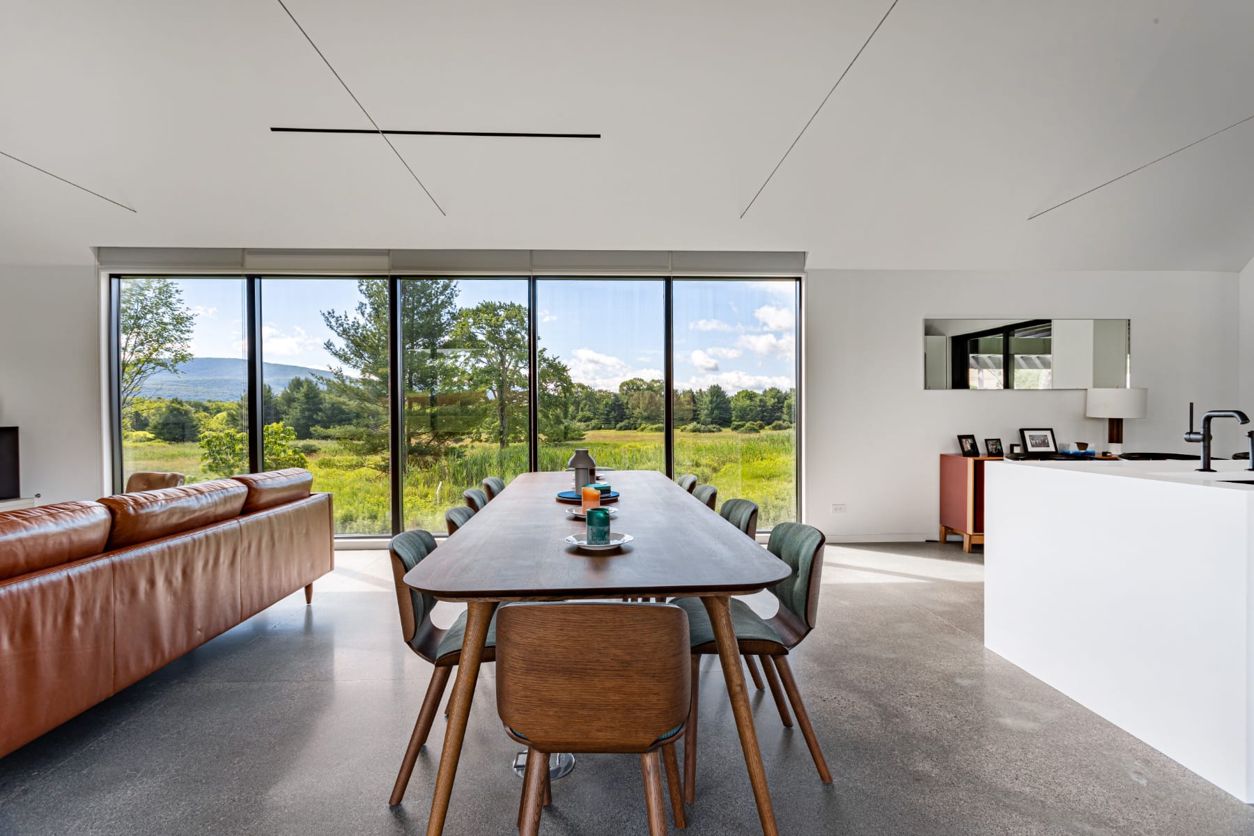 Modern dining area with a long wooden table and eight mid-century style chairs, set on polished concrete floors. A large floor-to-ceiling window provides a stunning view of a vibrant green landscape and distant mountains.