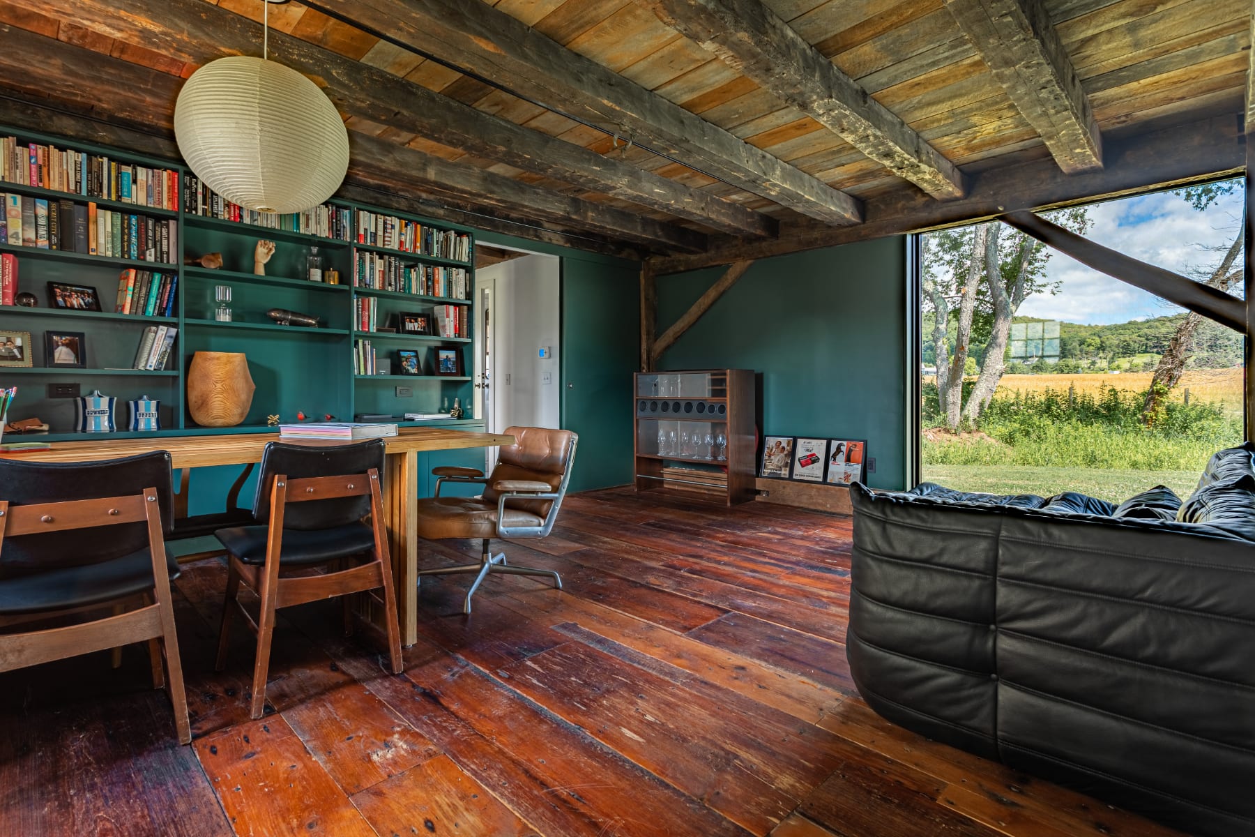 A rustic yet cozy room features dark green walls, exposed wooden ceiling beams, and rich wooden floorboards. A large bookshelf filled with books, a wooden desk with chairs, and a black leather sofa create a comfortable study or lounge space.