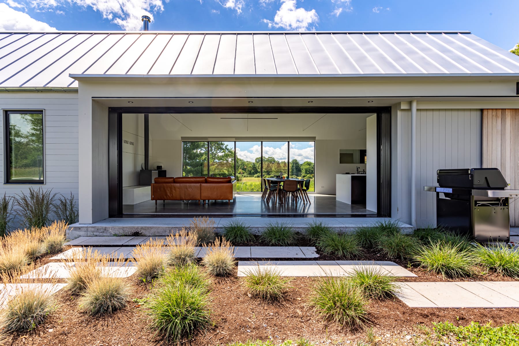 The modern rear exterior of the house features a white metal roof and light siding, with a wide opening revealing an open-concept living and dining area inside. A concrete path with decorative grasses leads to the entrance, and a BBQ grill is visible on the right.
