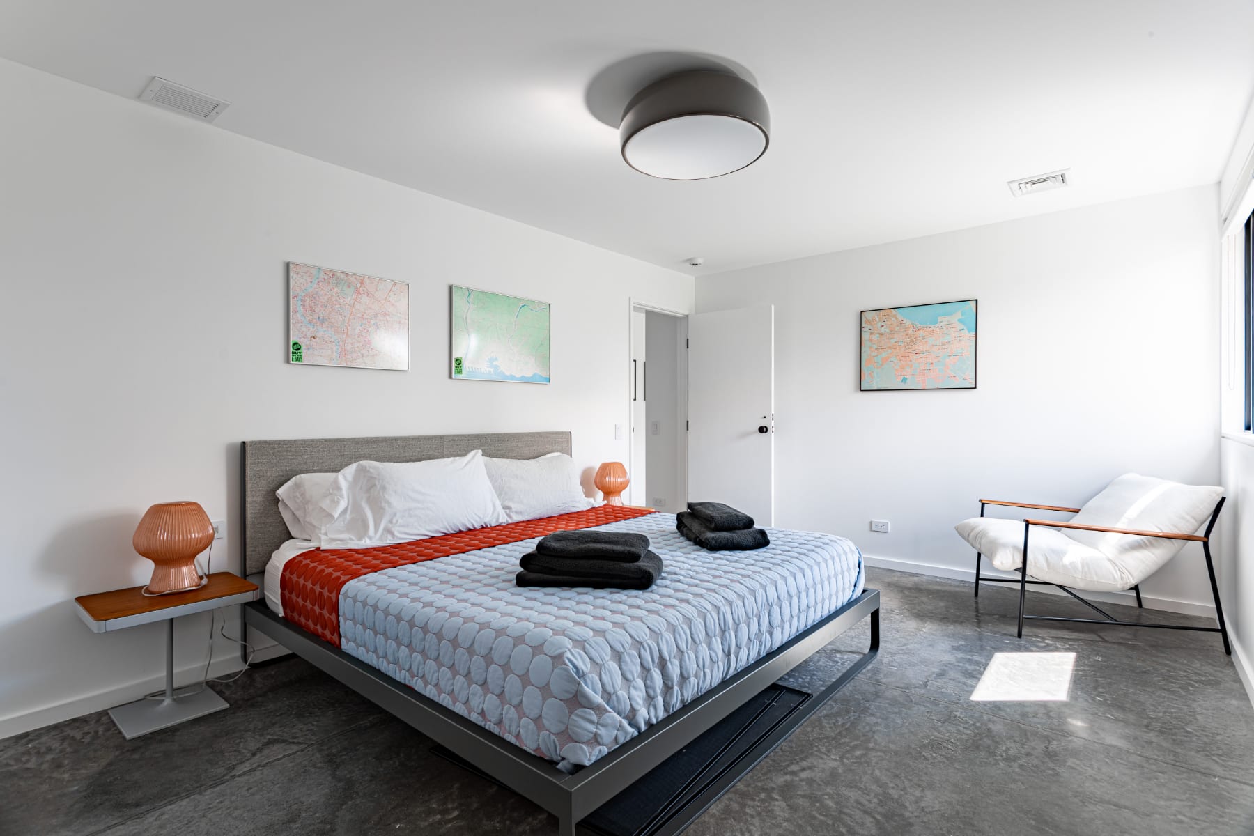 Modern bedroom featuring a king-sized bed with a grey upholstered headboard, light blue and orange bedding, and polished concrete floors. Two orange glass lamps sit on minimalist side tables, and framed maps decorate the white walls.